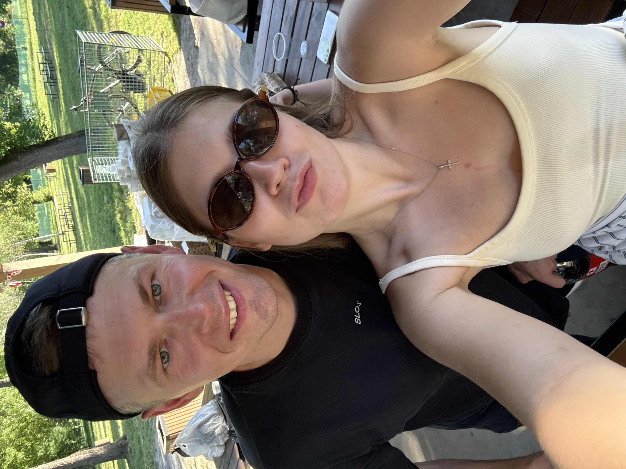 The image shows a man and a woman posing for a selfie. The man is wearing a black t-shirt and a black baseball cap. The woman is wearing a light-colored tank top and sunglasses. She also has a cross necklace. They are sitting outside at a wooden table. There is a green grassy area with some trees in the background. A bicycle in a cage can be seen in the background. Based on the background, the location is likely in a park or outdoor recreational area within the Cukarica Urban Municipality, Serbia.