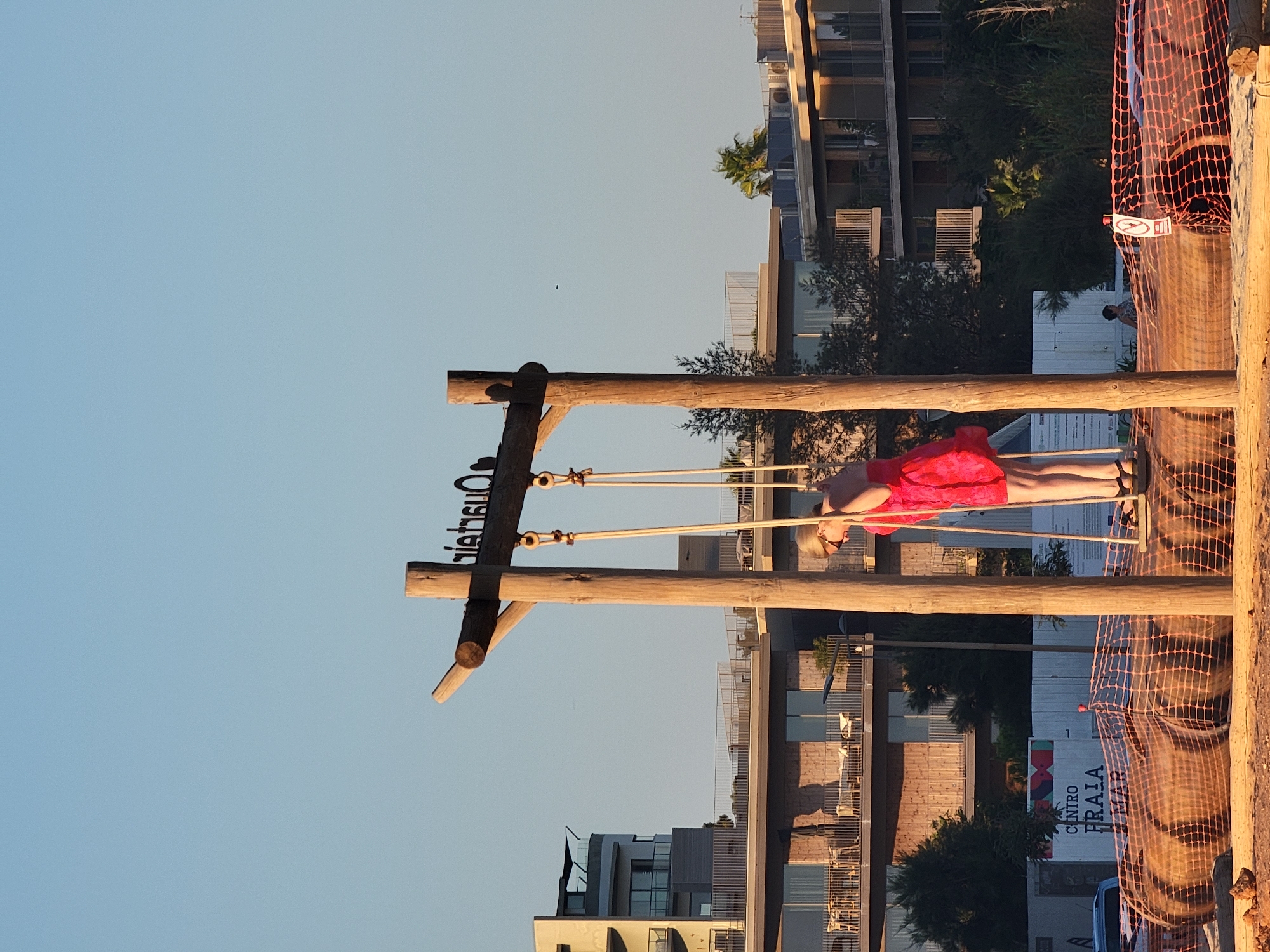 An adult female wearing a bright pink dress is standing on a swing set in Quarteira, Portugal. The swing set is made of natural wood and has two ropes suspending the seat. The woman is looking down and appears to be holding onto the ropes. In the background, there are modern multi-story buildings with balconies and a clear blue sky. The foreground shows a sandy area with construction netting and large cylindrical objects wrapped in netting, suggesting ongoing development or renovation. The overall scene captures a moment of leisure against a backdrop of contemporary architecture and construction.