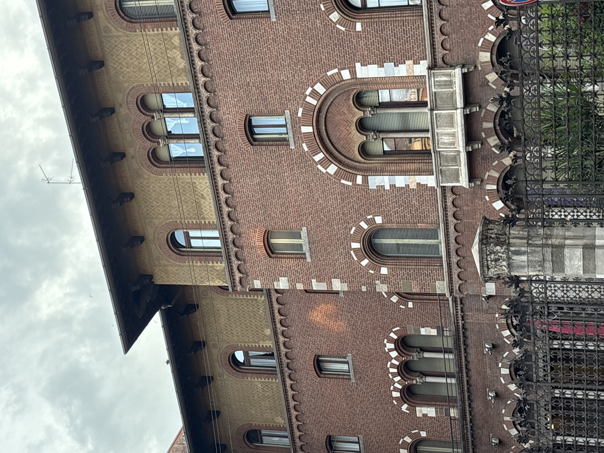 The image shows a portion of a brick building in Milan, Italy. The building features arched windows with white accents and a decorative cornice. A wrought-iron fence partially obscures the lower right corner of the building. The sky is cloudy.