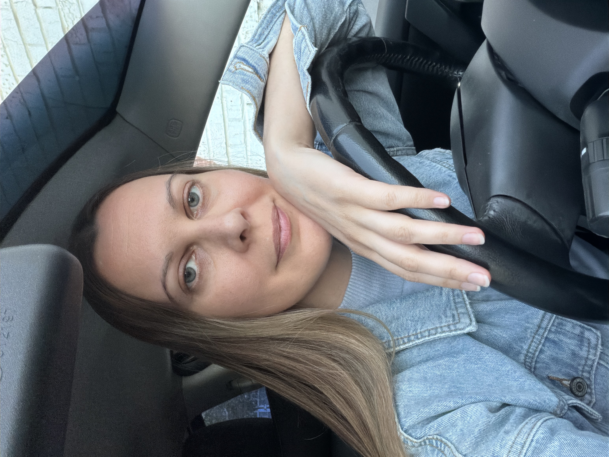 A woman with light brown hair and light eyes is posing in what appears to be the driver's seat of a car. She is wearing a light blue denim jacket over a lighter blue top. Her left hand is resting on the steering wheel. The image appears to be taken from inside the vehicle. The location of the image cannot be confirmed as Izhevsk, Russia, since there is no discernible context of such place.