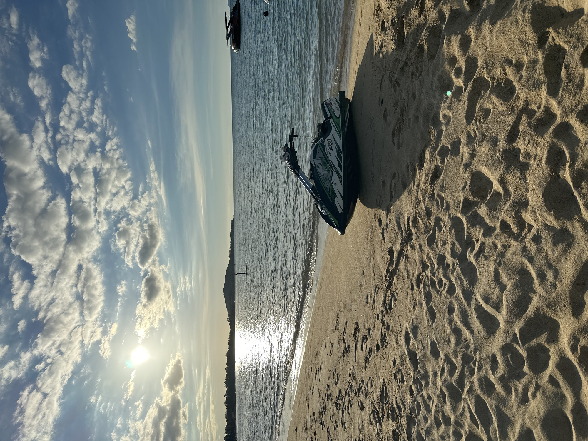 The image depicts a serene beach scene at Baan Mae Nam, Thailand, likely in the late afternoon given the warm, angled sunlight casting long shadows across the sand. The sky is a mix of blue and white, with scattered, wispy clouds that are illuminated by the sun, creating a sense of a pleasant, clear day.
The foreground is dominated by a sandy beach, marked with numerous footprints, indicating recent activity. A dark-colored jet ski, with green accents and some visible white text that appears to read "jet ski" and other brand-specific markings, rests on the sand near the water's edge.
The middle ground features the gentle lapping of waves on the shore, with the sun's reflection shimmering on the water's surface, creating a bright, almost dazzling effect. Further out in the sea, a few small boats or buoys are visible, hinting at marine activity or leisure. In the distance, a hazy, mountainous silhouette suggests the island's topography.
The overall mood conveyed is one of tranquility and relaxation. The lighting and natural elements suggest a peaceful moment on a tropical beach, ideal for water sports or simply enjoying the coastal ambiance. There are no people visible in the frame.