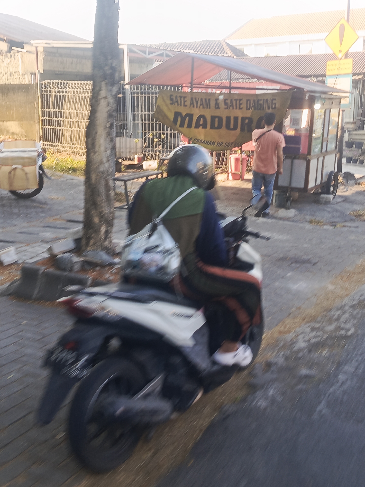 The image depicts a street scene in Surabaya, Indonesia, featuring a food stall selling "SATE AYAM & SATE DAGING MADURA." A person wearing a green jacket and a helmet is seated on a motorcycle in the foreground, facing away from the camera. The motorcycle is white and black. Behind the person on the motorcycle, a man in a peach-colored shirt and blue jeans stands near the food stall, appearing to be a vendor or customer. The food stall has a red awning and various items displayed within it. The environment includes a paved sidewalk, a tree, and buildings in the background. The image has motion blur, suggesting movement of the camera or subjects.