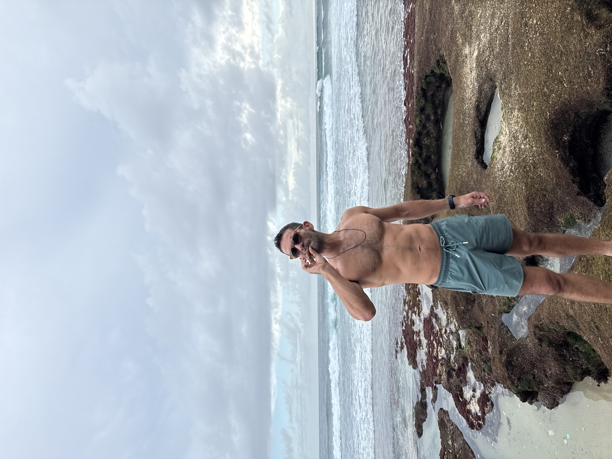 The image depicts a fair-skinned, shirtless man wearing dark sunglasses and green swim trunks standing on a rocky beach at Pecatu, Indonesia. He has a thin necklace and a watch on his left wrist, and appears to be holding something small between his fingers near his mouth. Behind him is the ocean with waves breaking onto the shore. The sky is overcast with dense clouds. The rocks in the foreground are covered with what appears to be reddish-brown algae.