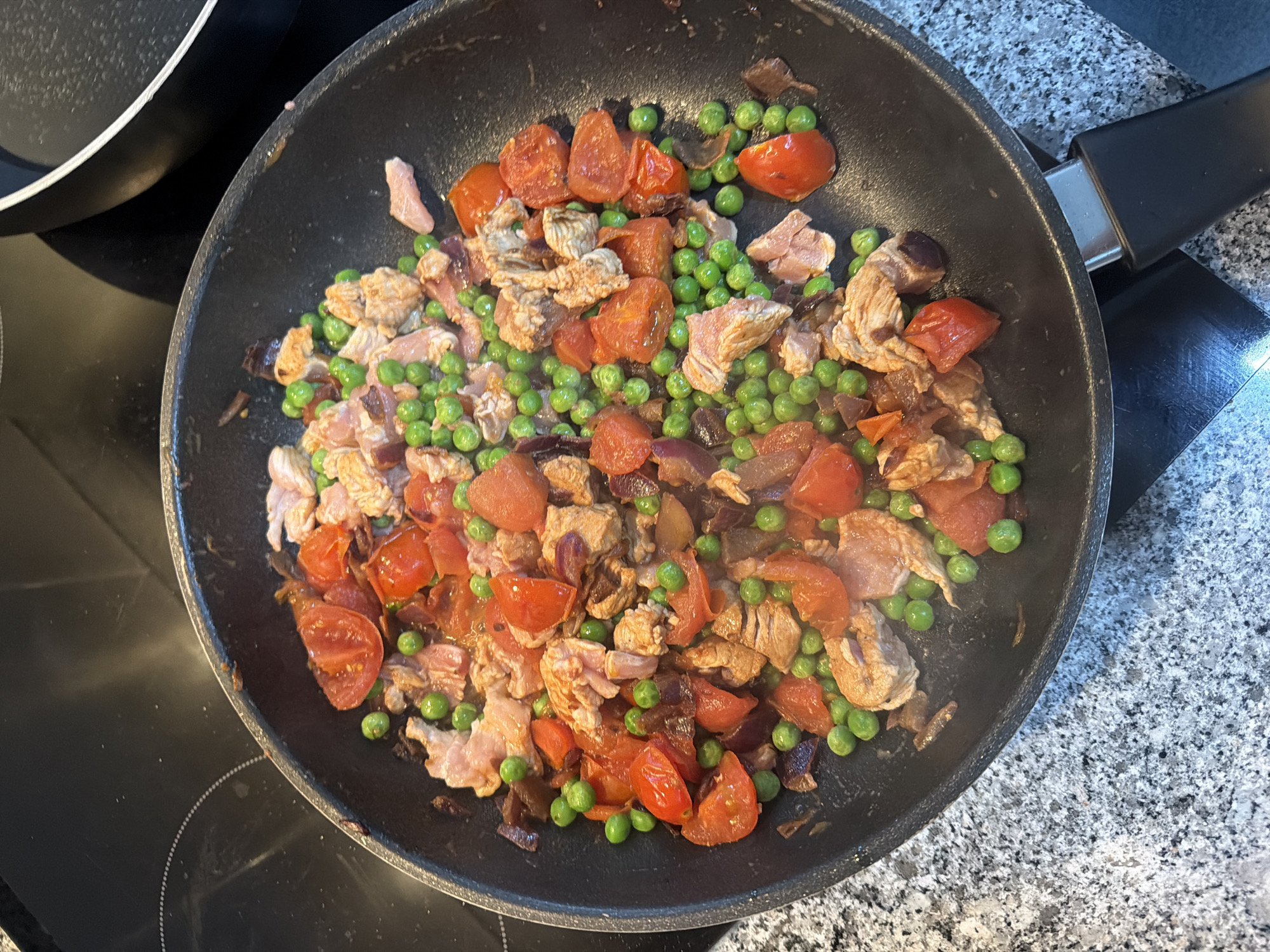 A top-down, medium shot shows a black frying pan on a black stovetop. Inside the pan is a mixture of cooked ingredients: cubed tomatoes, green peas, chopped meat (possibly chicken or pork), and diced red onions. The pan sits slightly off-center on the stovetop. Part of another pan is visible on the upper left. The frying pan has a black handle extending to the right, resting on a countertop with a mottled gray pattern. The image suggests a domestic cooking environment, possibly in a residential kitchen. The specific location is Vila Real, Portugal, though no evidence of such is visible in the image.