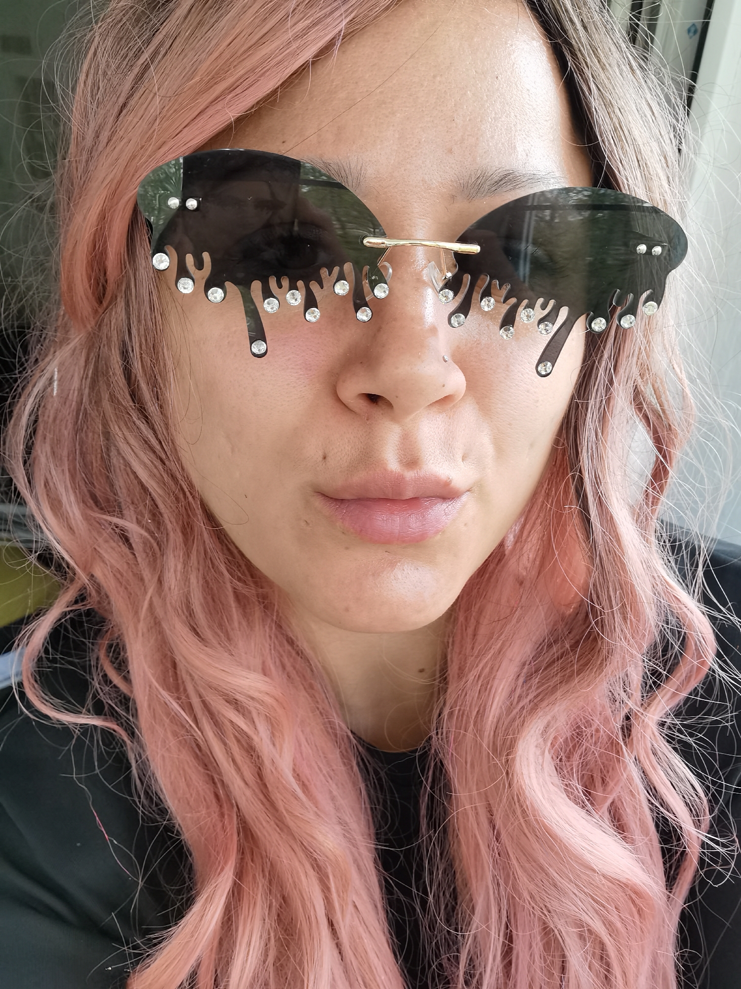 A close-up selfie shows a person with pink wavy hair and black sunglasses with a "dripping" design, each "drip" adorned with a small jewel. The person has a nose piercing and fair skin. The background is slightly out of focus, revealing a partially visible window or doorway with muted light. The person wears a black shirt or top. The specific location in Chaikovsky, Russia cannot be determined from the visual information provided.