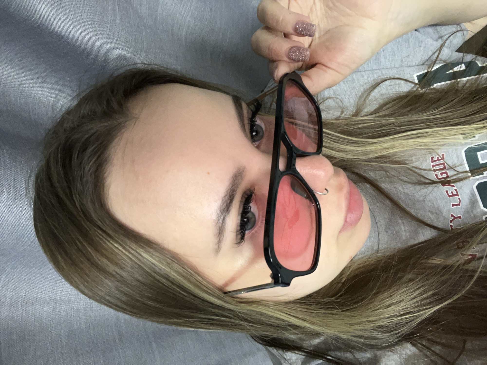 A young woman lies on a gray surface, possibly a bed or couch, wearing a gray sweatshirt. She is holding black-framed glasses with red lenses in front of her face. Her hair is light brown with blonde highlights. She has a nose piercing on her left nostril. The sweatshirt has a dark green and red graphic on it with the word "LEAGUE" visible. The background is a plain gray material. There's no clear indication this image was taken in Krasnodar, Russia.