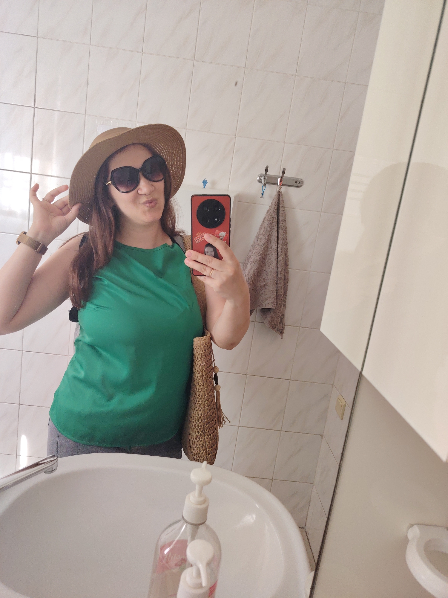 A woman is taking a mirror selfie in a bathroom. She is wearing a green sleeveless top, grey pants, a straw hat, and large sunglasses. She is holding her hat with her left hand and her phone with her right hand, as if to take a picture. She is also carrying a straw bag over her shoulder. The bathroom has white tiled walls and a white sink with a faucet and a soap dispenser. A towel is hanging on a hook on the wall. The woman is pouting her lips. The image does not provide any information about the city of Rostov-on-Don, Russia.