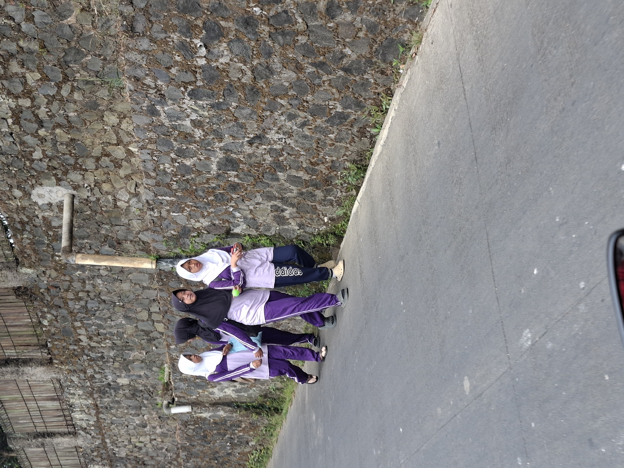 In Balekambang, Indonesia, four young people, likely students wearing purple uniforms and hijabs, walk along an asphalt road. They are positioned against a textured stone wall that forms the backdrop of the scene. The wall appears to be constructed from irregular stones, with some vegetation growing along its base and on its surface. A large, light-colored pipe runs horizontally across the upper portion of the wall. The road surface is gray asphalt, with visible seams and some debris. The lighting suggests it is daytime, with shadows cast on the road. The subjects are walking in the same direction, away from the viewer, with some of them holding small objects.