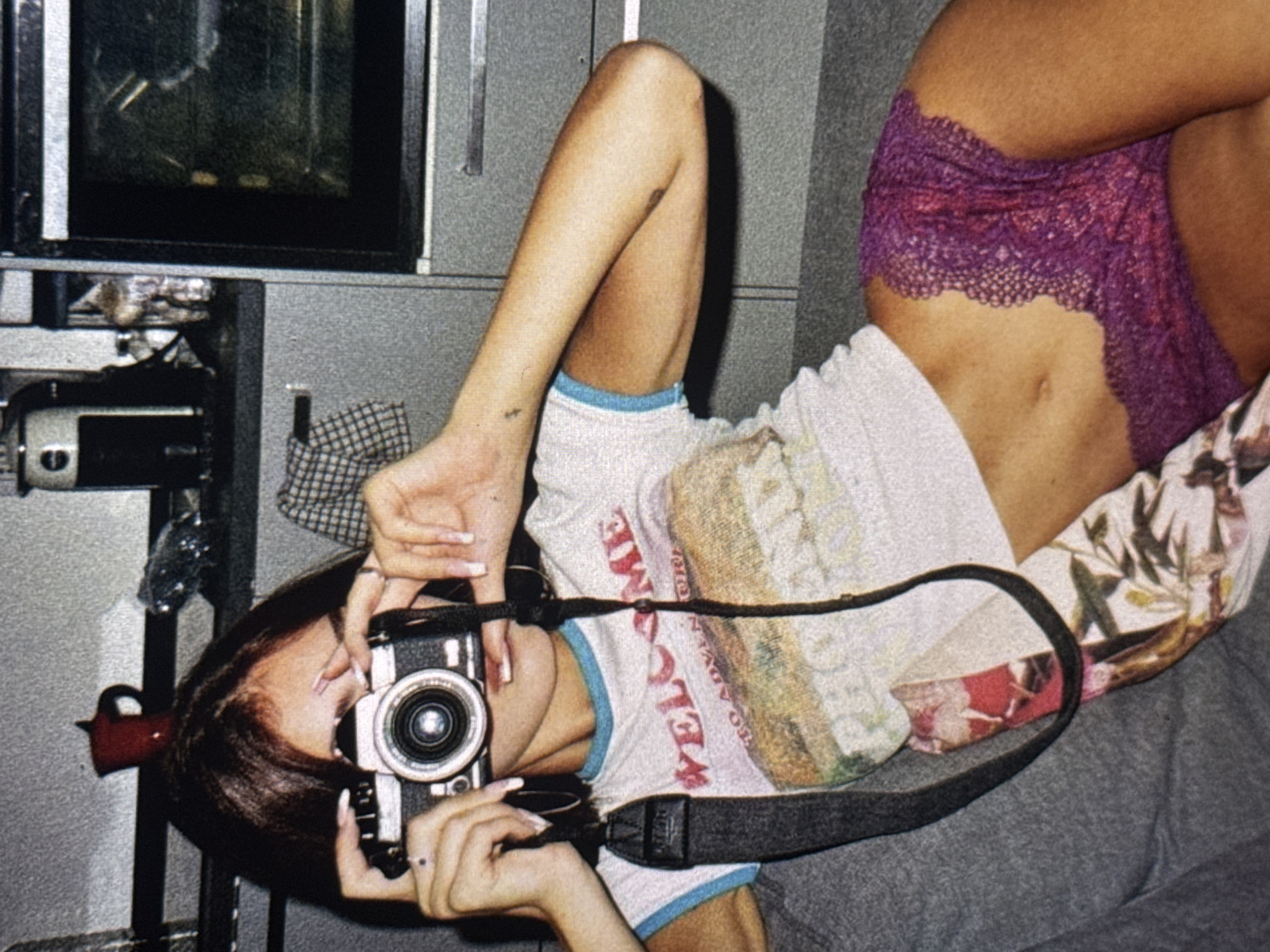A young woman with dark hair and light skin is pictured in a medium shot, holding a vintage camera up to her face as if taking a photograph. She is wearing a white t-shirt with a graphic design and blue trim, and purple lace underwear. The background appears to be a kitchen area with cabinets, an oven, and shelves holding various items, including a red mug and a checkered cloth. The overall lighting is dim, suggesting an indoor setting, possibly a home or a rented accommodation. There is no indication of the specific location in Kuta Selatan, Indonesia.