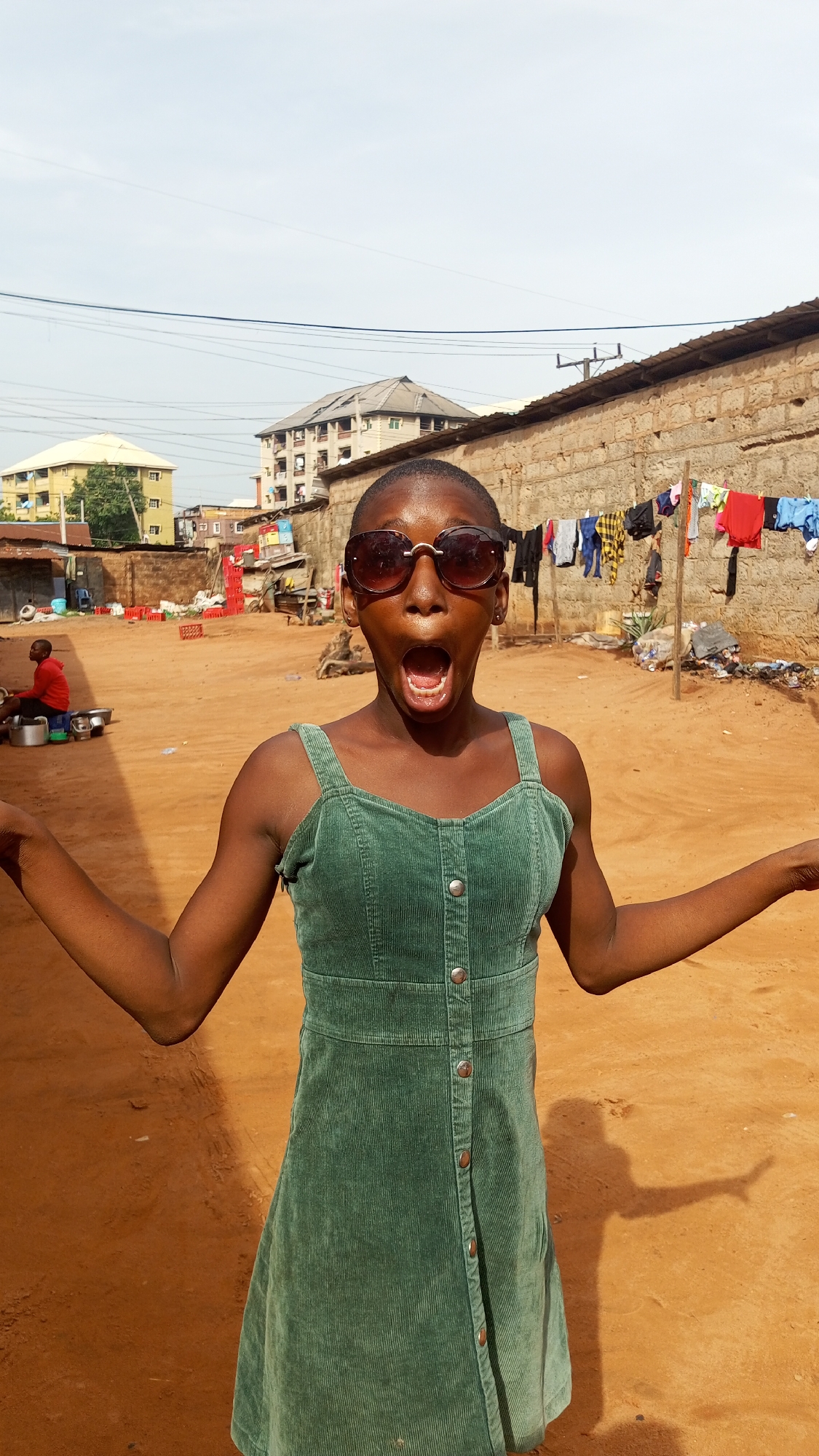 A young person stands with arms outstretched and mouth open in Nkpor, Nigeria. The person wears sunglasses and a sleeveless green dress with button closures down the front. Behind them is a dusty, sunlit area with buildings, a stone wall, and laundry drying on a line. A person in a red shirt sits on the ground near metal pots. Crates and other objects are scattered around the sandy ground. Overhead electrical wires stretch across the sky.