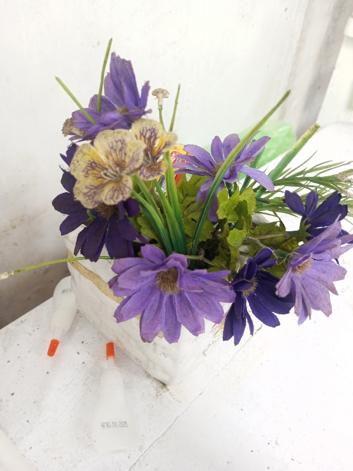 The scene, located in Ciherang, Indonesia, displays a white rectangular planter holding an arrangement of artificial flowers. The floral assortment comprises numerous purple and light purple daisy-like blossoms, intermixed with several beige and brown-striped flowers resembling pansies, along with artificial green leaves and stems. To the left of the planter, positioned on a textured, light-colored surface, are two small, translucent white bottles, each fitted with an orange dropper nozzle. One bottle bears the inscription "H 30.09.2025". The background consists of a plain, light-colored wall, with a subtle green object partially visible behind the flower arrangement.