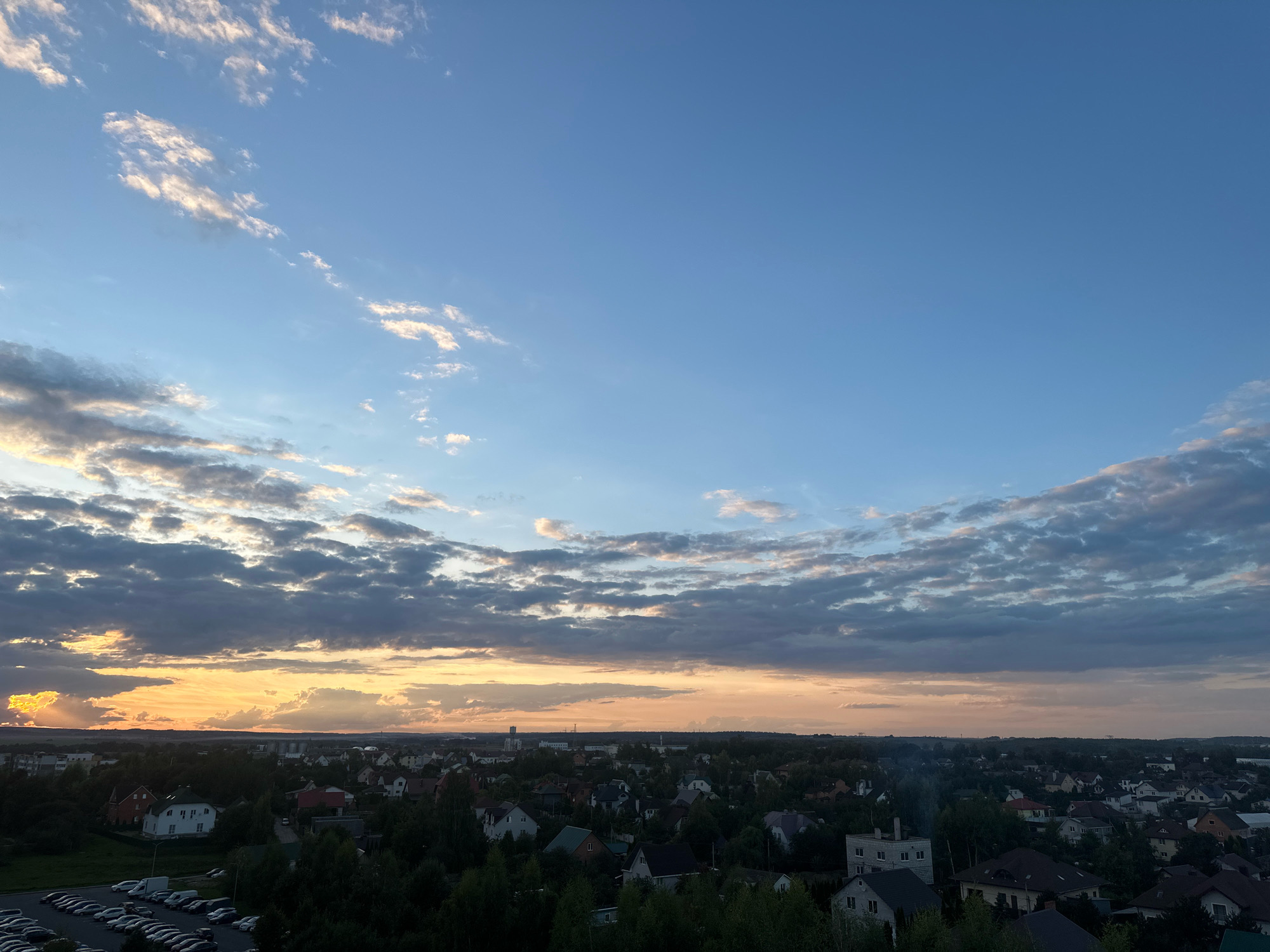 The image captures a panoramic view of a residential area in the vicinity of Шчомысліцкі сельскі Савет, Belarus, during what appears to be sunset. The sky is a gradient of blue, transitioning to hues of orange and yellow near the horizon, indicative of the sun setting. Scattered clouds, some tinged with the warm colors of sunset and others a darker gray, drift across the sky.
The landscape below consists of numerous houses with various roof styles and colors, interspersed with abundant green trees. In the foreground, a large parking lot is visible, filled with numerous cars parked in neat rows. A white van is also present in the parking lot. The residential buildings are a mix of single-family homes, with some appearing to be multi-story. A few industrial-looking structures are visible in the distance, near the horizon. The overall scene depicts a suburban or semi-rural setting within the Шчомысліцкі сельскі Савет region, bathed in the soft, warm light of dusk. There are no people visible in the image.