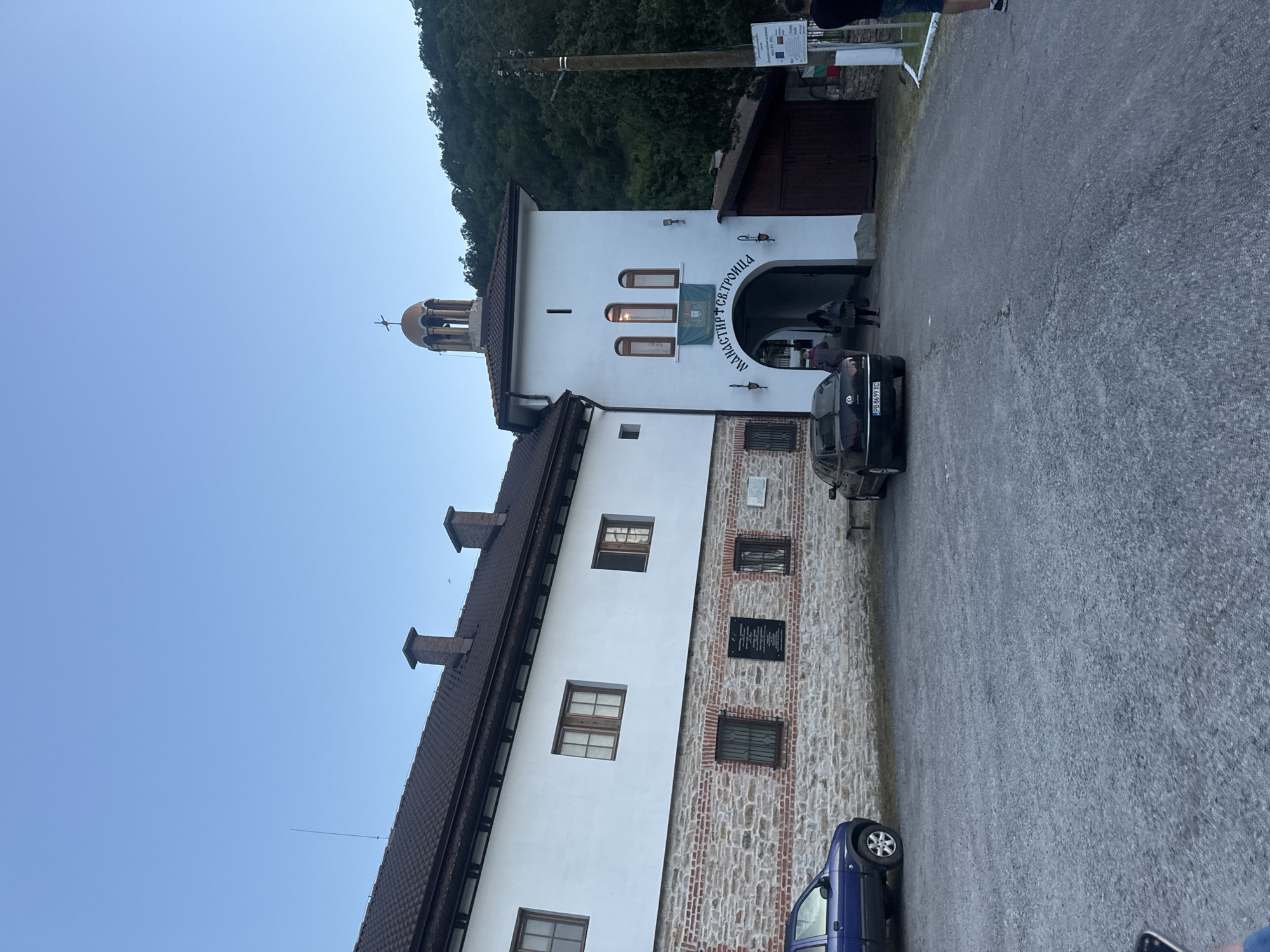 The image depicts the facade of the "St. Trinity Monastery" located in Mramor, Bulgaria. The building is primarily white, with a section built from stone. It features a bell tower, a dark brown tile roof, and several windows. A dark-colored car is parked in front of the arched entrance. Part of a blue car is visible in the foreground. The background includes a forested hillside. The sky is pale blue, indicating either early morning or late evening. A sign with text in Cyrillic script is visible above the entrance.