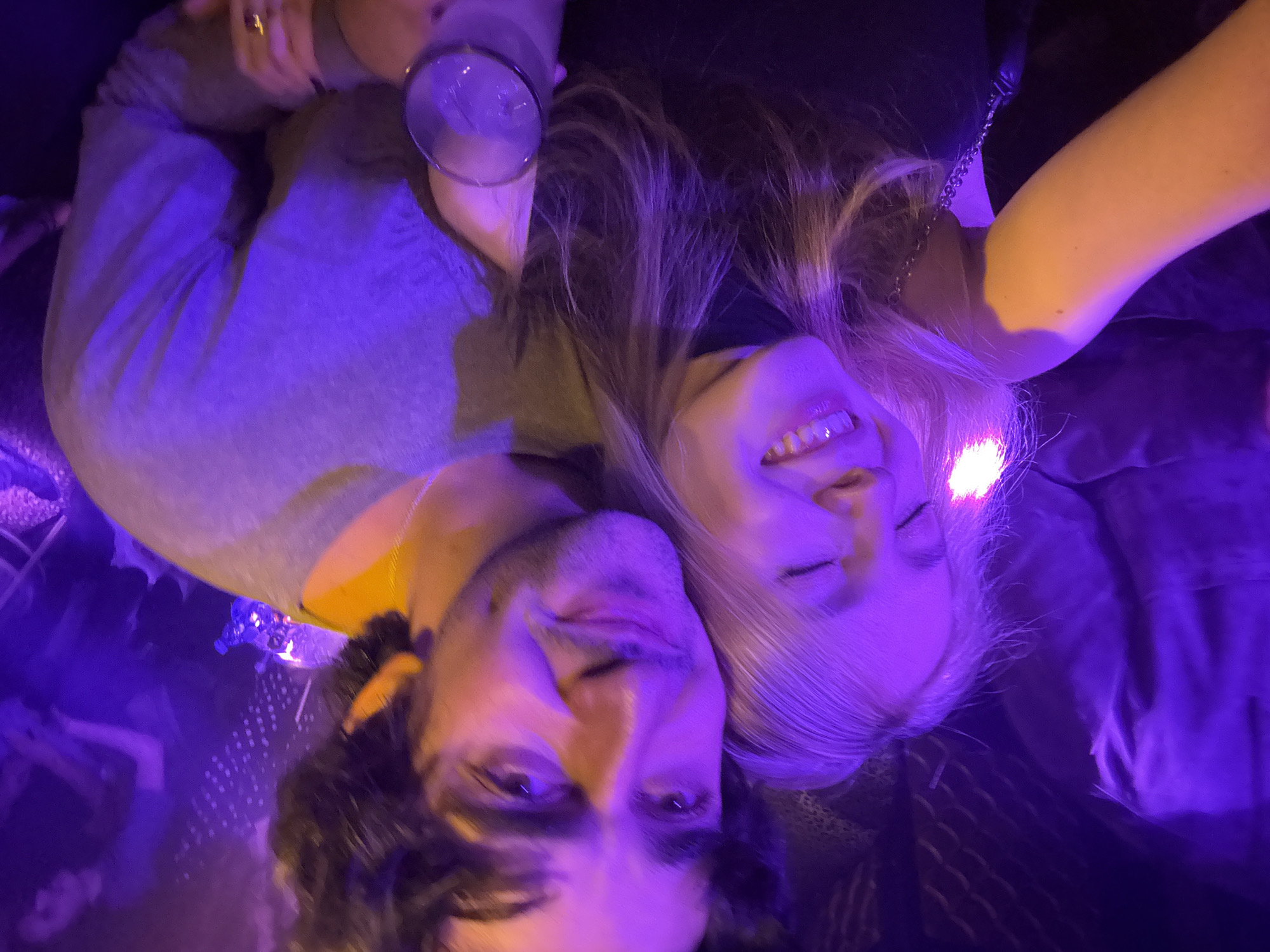 Two individuals are visible in the image, a man and a woman, bathed in purple light. The woman is fair-skinned with blonde hair. She is smiling and appears to have her eyes closed. The man has dark hair and a beard. The image is taken from an unusual angle, possibly upside down or sideways. A glass filled with liquid is visible in the upper left corner. The location appears to be a dimly lit indoor space, likely a bar or club, based on the lighting and casual attire of the subjects. The city is unknown.