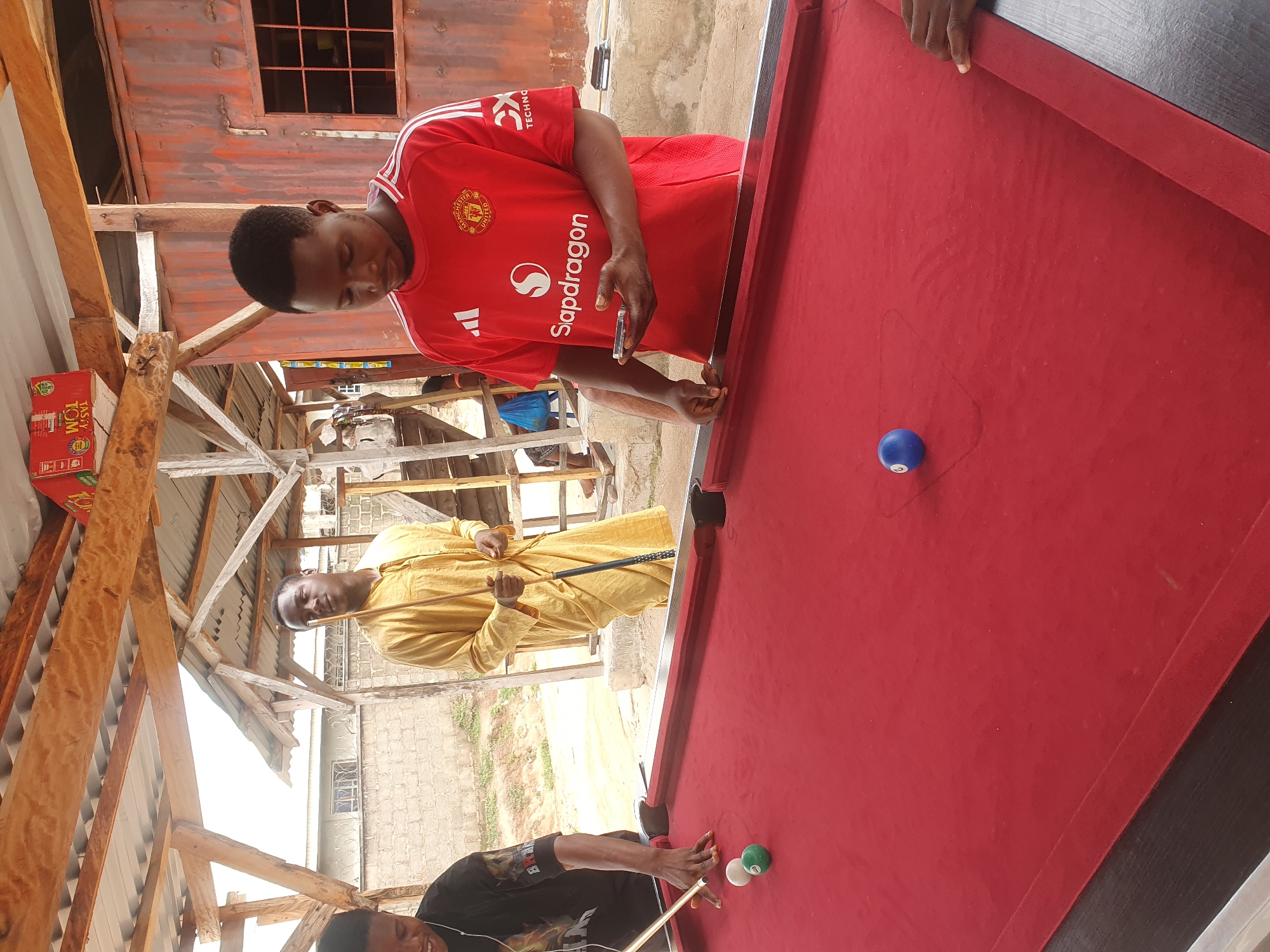 The image depicts an outdoor billiard scene in Lokoja, Nigeria. Three young men are present. One man, wearing a yellow traditional garment, stands holding a billiard cue. Another man, dressed in a black t-shirt, is in the process of taking a shot at a billiard table with a red felt surface. A third man, wearing a red Manchester United jersey with "Snapdragon" branding, stands to the right, holding a smartphone. The billiard table is set up under a rustic wooden and corrugated metal roof structure. In the background, a brick wall and a dilapidated building are visible. The lighting suggests it is daytime.