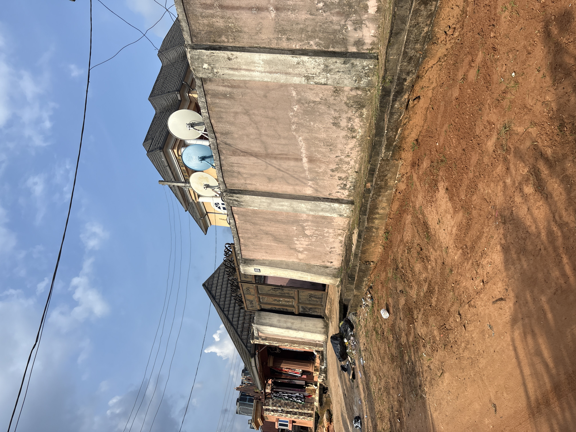 In Obosi, Nigeria, a dirt road leads past a high, pink, concrete wall topped with several satellite dishes. A large, ornate metal gate is visible in the wall, and to the left of the gate, a covered structure appears to be a shop or residence, with clothes hanging outside. The ground is a mixture of reddish-brown dirt and debris, including several black trash bags. The sky is blue with scattered white clouds.