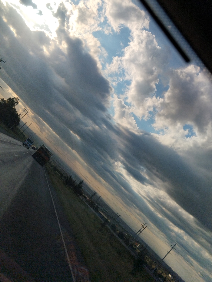 The image shows a view of a highway from a car window. The road is dark asphalt, with a white line down the center and other lanes to the left. There are several vehicles in the distance on the highway, including a truck with a yellow cab. The highway is lined with green grass, with trees and shrubs on the side of the road. In the background, there are multiple power lines and a distant cityscape that extends towards the horizon. The sky is cloudy with a few areas of blue visible, and there are some bright rays of sunlight peeking through the clouds. The image is taken from a location on the Athi River Road in Athi River, Kenya at latitude: -1.475, longitude: 37.034.