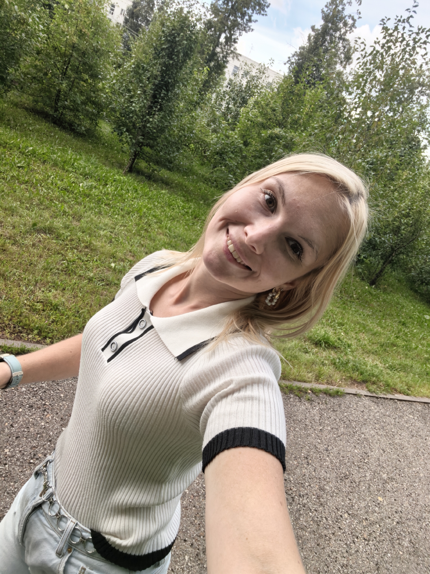 A young Caucasian woman with blonde hair, brown eyes, and light skin is taking a selfie outdoors. She is wearing a white ribbed polo shirt with black trim and buttons, light-wash denim jeans with a silver ring belt, and a light blue wristwatch. She is smiling and looking at the camera. The background consists of a grassy slope with trees and a paved path. A building and trees are visible in the distance under a cloudy sky. The image appears to have been taken in a park or green space within the city of Sterlitamak, Russia.