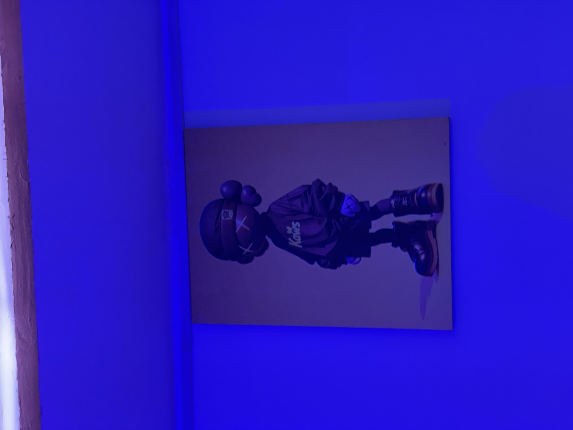 A framed artwork of the KAWS Companion character is displayed on a wall under blue lighting. The artwork depicts the character from the waist down, wearing black shorts, a black long-sleeved shirt with "KAWS" written on it, and black boots. The character has distinctive X's for eyes and round ears. The blue lighting casts a vibrant glow on the scene, creating a moody atmosphere. The image does not provide information about the specific location within the city of Durimi, Nigeria.