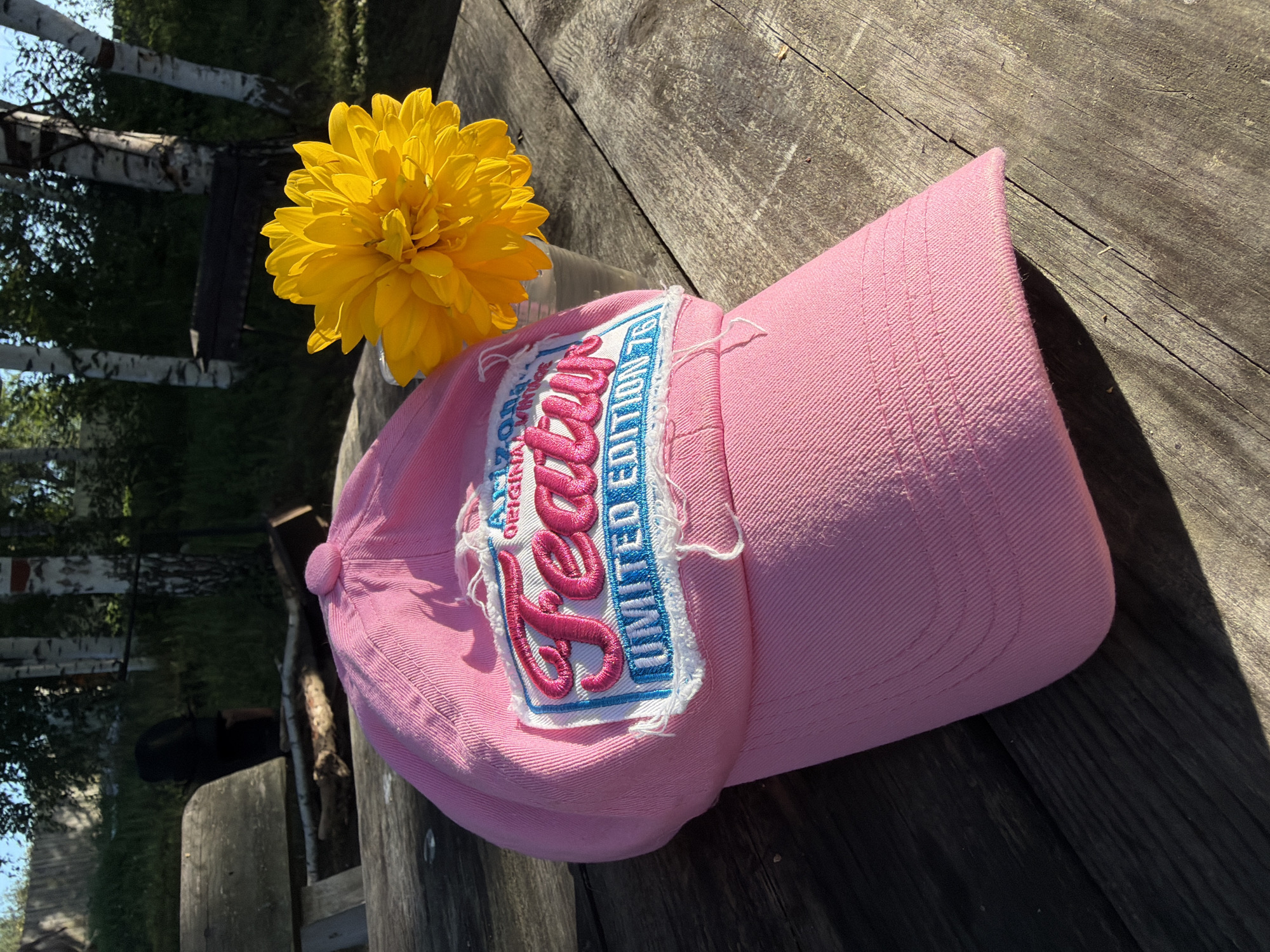 A pink baseball cap with a distressed patch is placed on a weathered wooden picnic table outdoors. The patch on the cap reads "ARIZONA ORIGINAL VINTAGE" in a blue rectangle with pink lettering that says "Featu" and "LIMITED EDITION 76". Next to the cap, a bright yellow flower in a clear plastic cup sits on the table. In the background, there are trees, including birch trees, and a wooden bench. The scene appears to be in a natural, outdoor setting, possibly a park or a backyard. This image was taken in Zonovo, Russia.