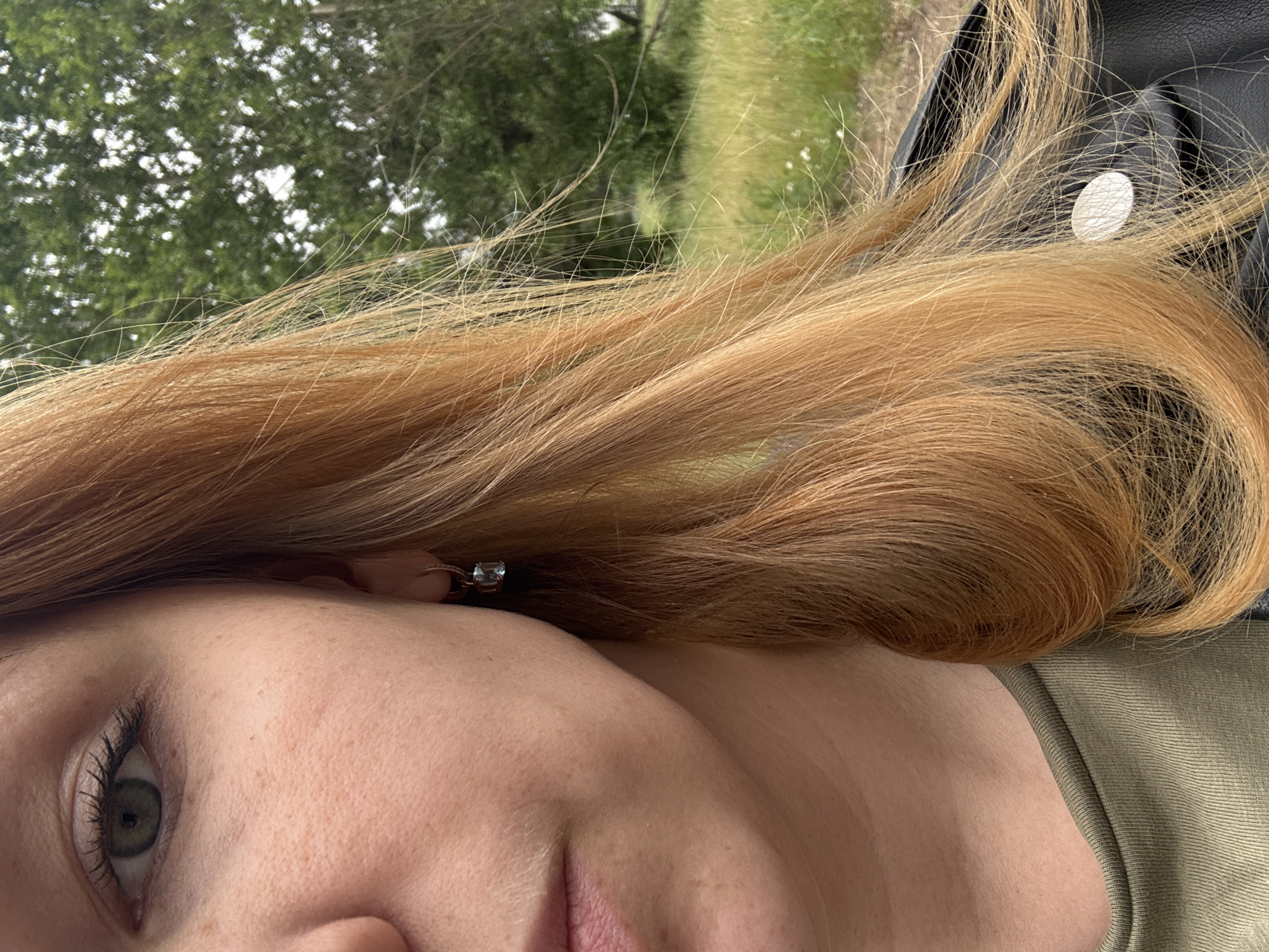 The image shows a close-up, cropped view of a person's face, focusing on their left eye, cheek, and mouth. They have fair skin with freckles and green eyes with visible eyelashes. A small, light blue earring with a rectangular stone is visible on their left ear. Their hair is a prominent feature, with strands of light brown or auburn hair cascading across the frame. The background is blurred, suggesting an outdoor environment with green trees and vegetation, possibly a park or natural area. The person is wearing a green t-shirt and a black leather jacket. The scene does not provide any specific clues about the city of Заволжье, Russia.