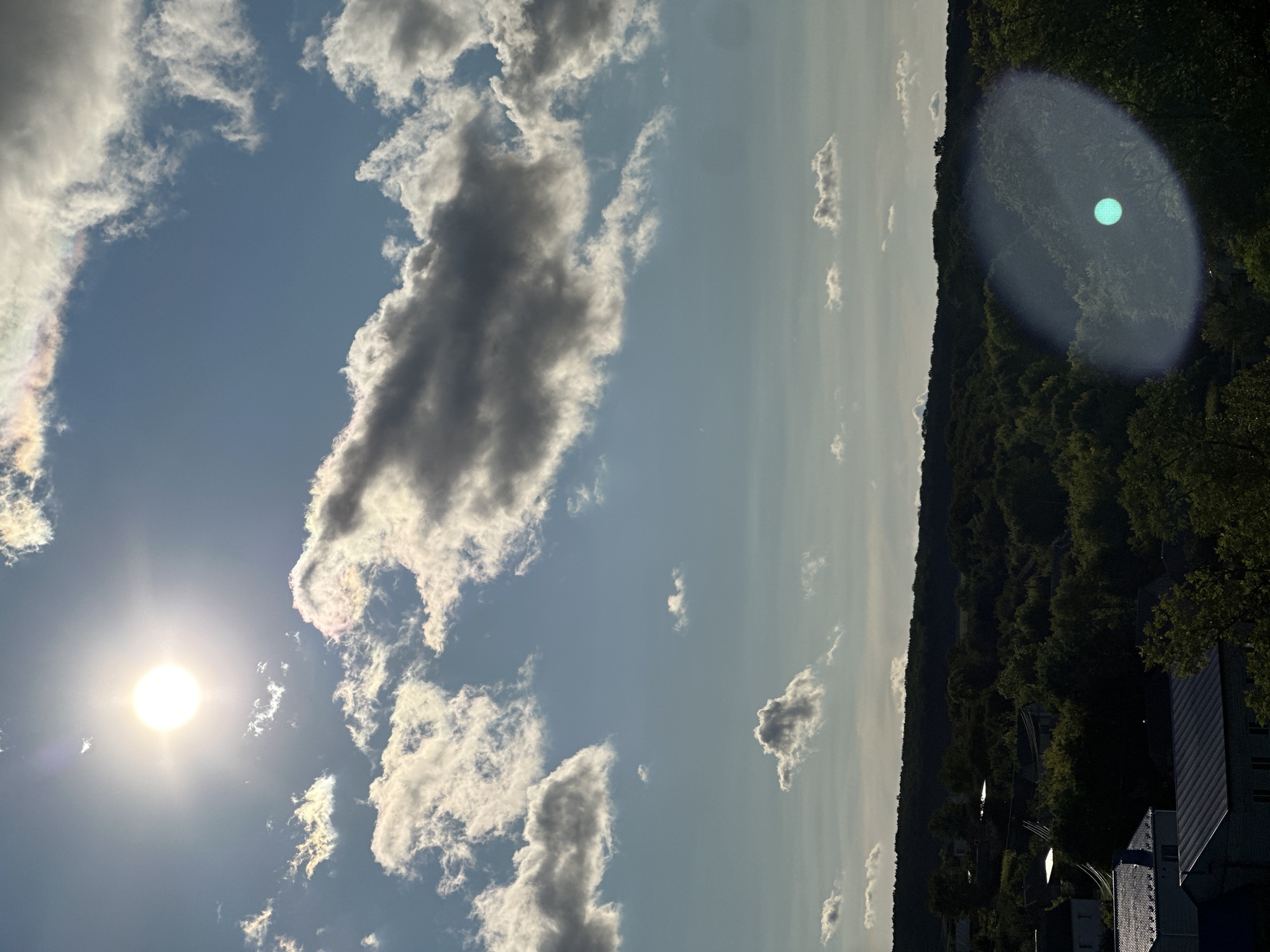The image depicts a sun-drenched sky above a densely wooded hillside with scattered houses in Makiv, Ukraine. The sun, positioned in the upper left quadrant, casts a bright light, creating lens flare artifacts in the lower right. Cumulus clouds are scattered across the blue sky, with some exhibiting iridescence. The hillside below is covered in a dark green forest, with glimpses of houses and rooftops visible through the trees. The overall scene suggests a rural or semi-rural setting during daytime, likely late afternoon given the angle of the sun and the shadows.