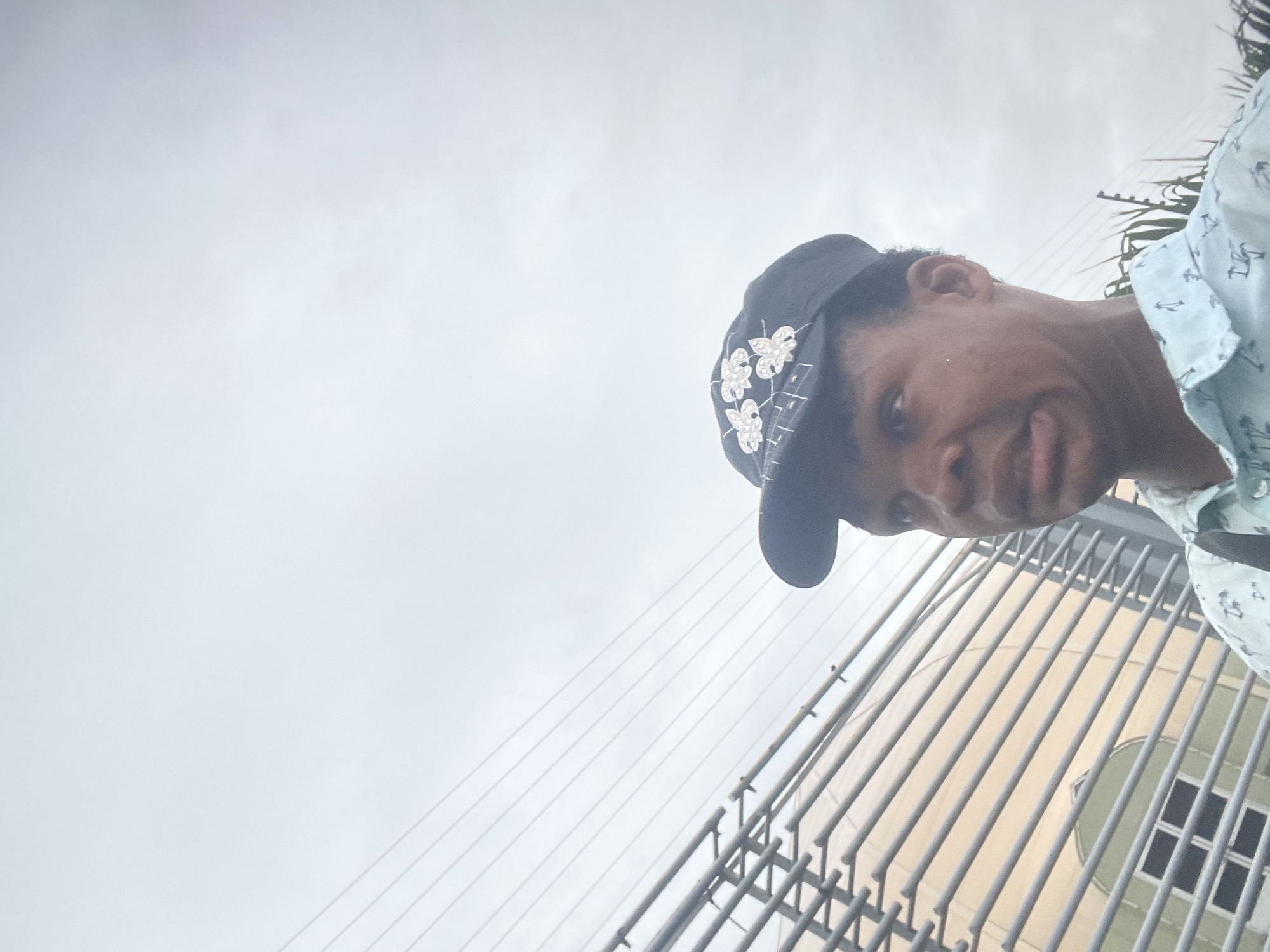 In Igboefon, Nigeria, a close-up shot captures a young Black person wearing a black baseball cap adorned with white floral appliques. The person is dressed in a light blue collared shirt featuring a repeating pattern of palm trees and anchors. They are looking off to the left of the frame with a slightly distressed expression. In the background, horizontal metallic bars form a fence or railing, behind which a building with a light-colored facade and a section of green-tinted windows is visible. Overhead, a network of power lines stretches across an overcast sky. The composition is tight, focusing on the individual's face and upper torso, with the urban background providing context.