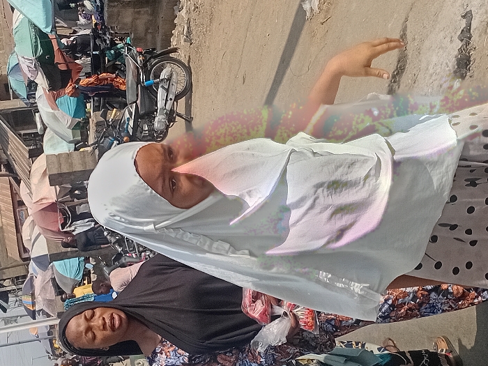 Two women pose in a bustling outdoor market. One woman, wearing a black hijab, looks directly at the camera with a slightly open mouth. Another woman, partially visible, is draped in a white hijab. A hand reaches out in the background toward a weathered wall. Motorbikes, stalls, and other shoppers create a chaotic market scene in the background. Umbrellas provide shade to the market stalls. The scene suggests a lively marketplace atmosphere.