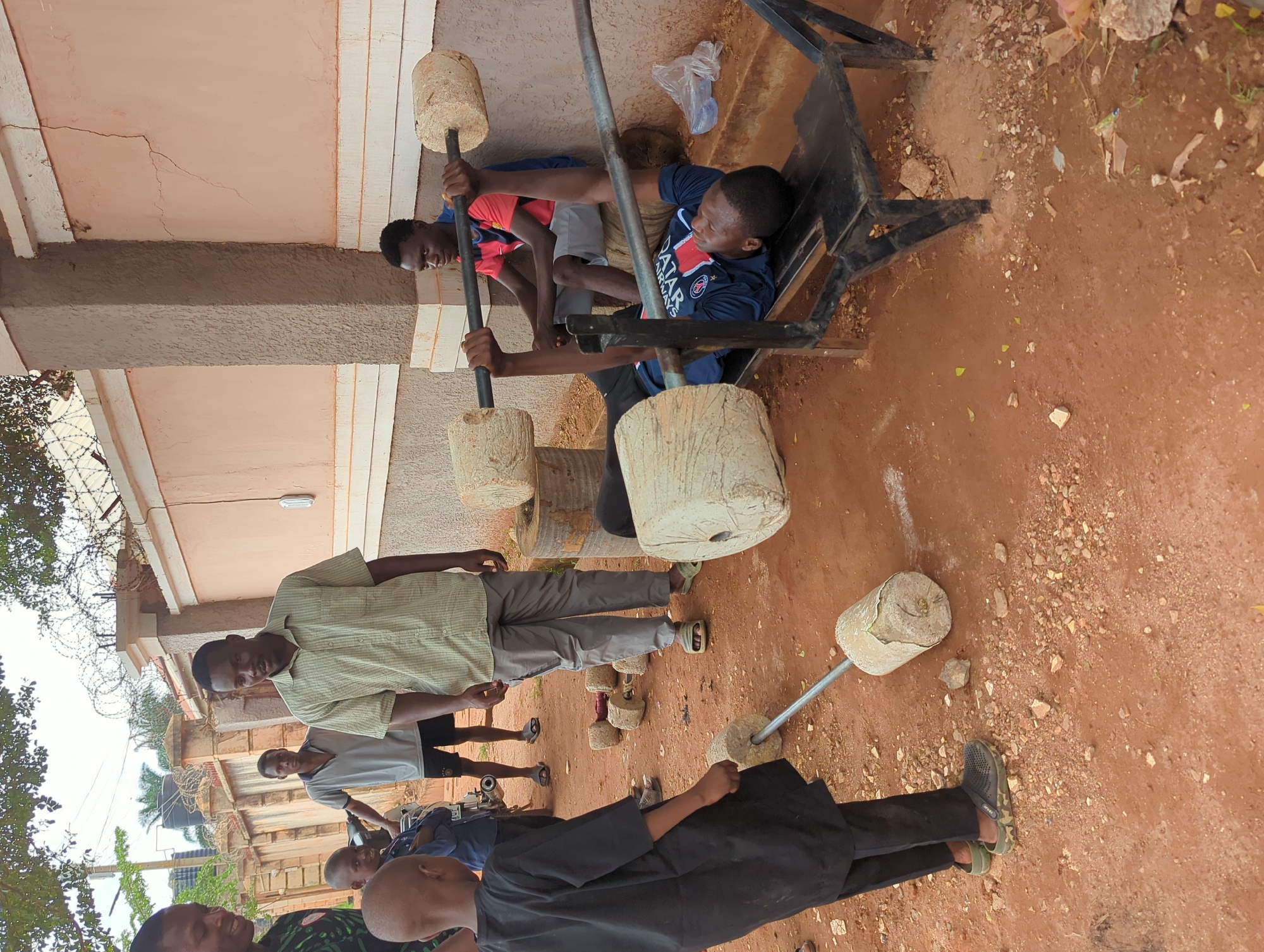 This image captures an outdoor scene in Unguwar Rimi, Nigeria, depicting a group of young men and boys engaged in physical activities and socializing.
In the foreground and midground, two young men are actively lifting weights. One young man, wearing a dark blue sports shirt with "QATAR AIRWAYS" and a PSG (Paris Saint-Germain) logo, is lying on a makeshift metal weight bench, pressing a barbell. The barbell is constructed from a metal rod with cylindrical concrete or stone weights at each end. Another young man stands nearby, holding a similar homemade barbell, ready to lift.
Several other individuals are present in the scene. A man in a light green plaid shirt and dark trousers stands observing, looking towards the camera. Further back, another person in a light grey shirt stands near a small motorcycle. Other figures are partially visible in the background, including boys.
The setting is an open area with reddish-brown dirt ground, scattered with small rocks and debris. The exercise takes place in front of a building with light-colored walls and a darker, textured lower section. Barbed wire is visible along a fence or wall in the background, suggesting an enclosed area. Some sparse greenery and trees are also visible.
The scene takes place during the day, under natural, diffused lighting. The notable detail is the improvised nature of the gym equipment, highlighting resourcefulness in creating fitness tools from available materials. The atmosphere is casual, portraying a communal fitness and social gathering.