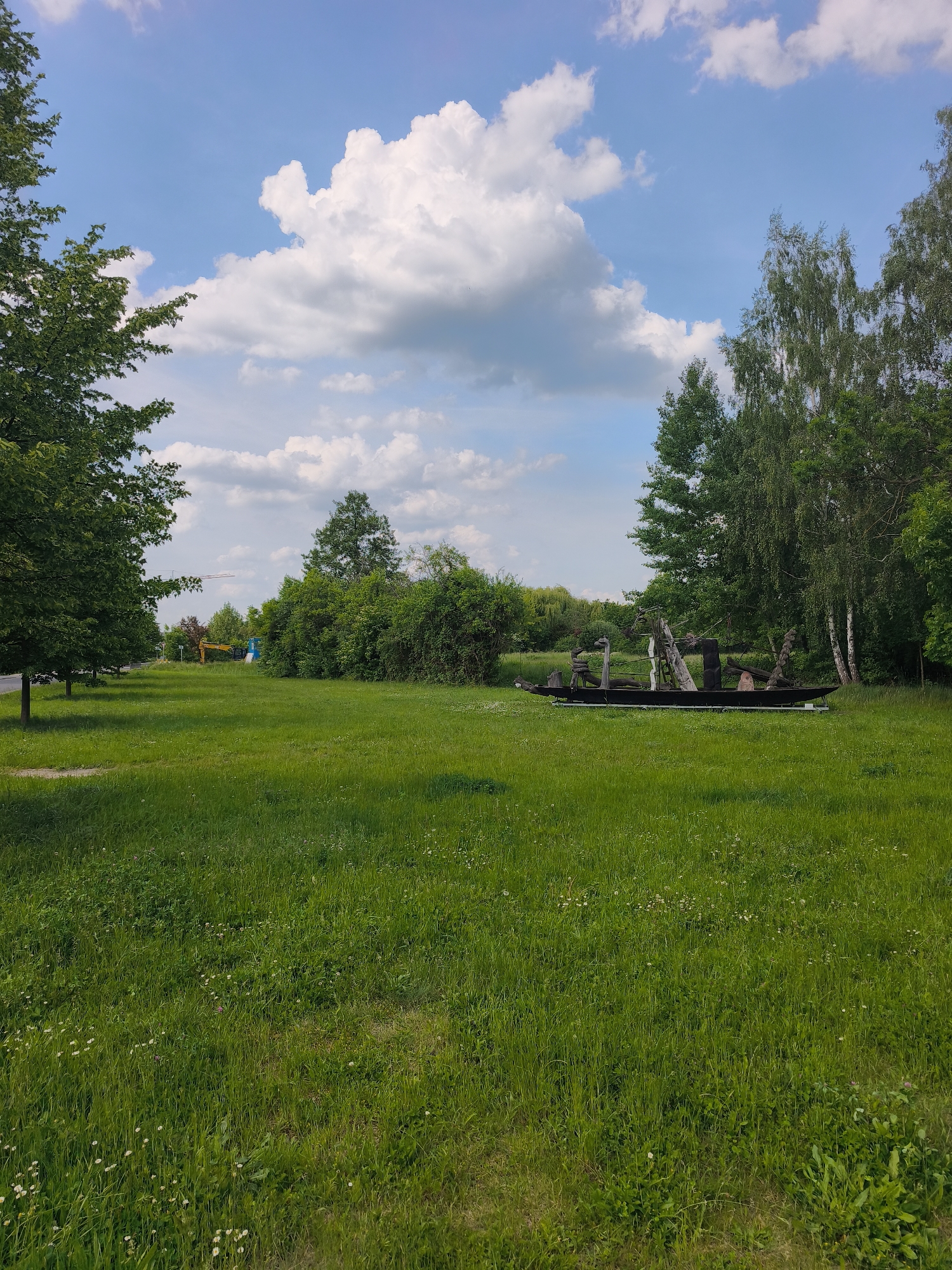 A daytime shot captures a grassy field bordered by trees under a partly cloudy sky. On the right, a cluster of birch trees and other greenery forms a natural barrier. In the middle of the field is an art installation resembling a boat constructed from dark wood and various wooden elements. The sky is blue with large, puffy white clouds. A line of trees and a road are visible in the distance on the left side of the image, with construction equipment subtly present in the background. The location is Unknown, Unknown.