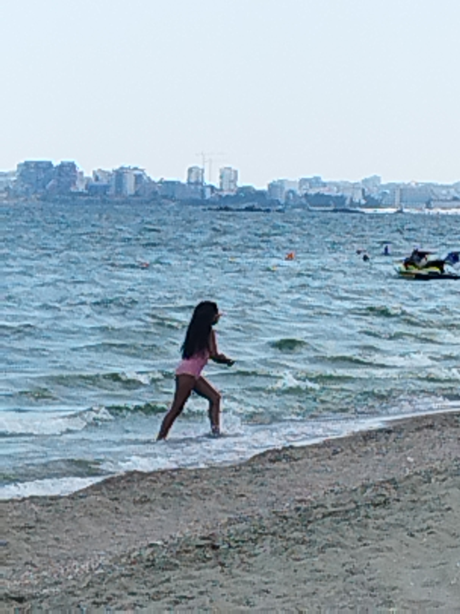 A young girl with dark hair is walking in the shallow water at the edge of a sandy beach. She is wearing a pink swimsuit. The sea is choppy with moderate waves. In the background, a city skyline with buildings and a crane can be seen across the water. A jet ski is visible further out in the sea. The sky is overcast, suggesting it might be a cloudy day. The scene appears to be during daylight hours. The overall impression is a casual day at the beach.