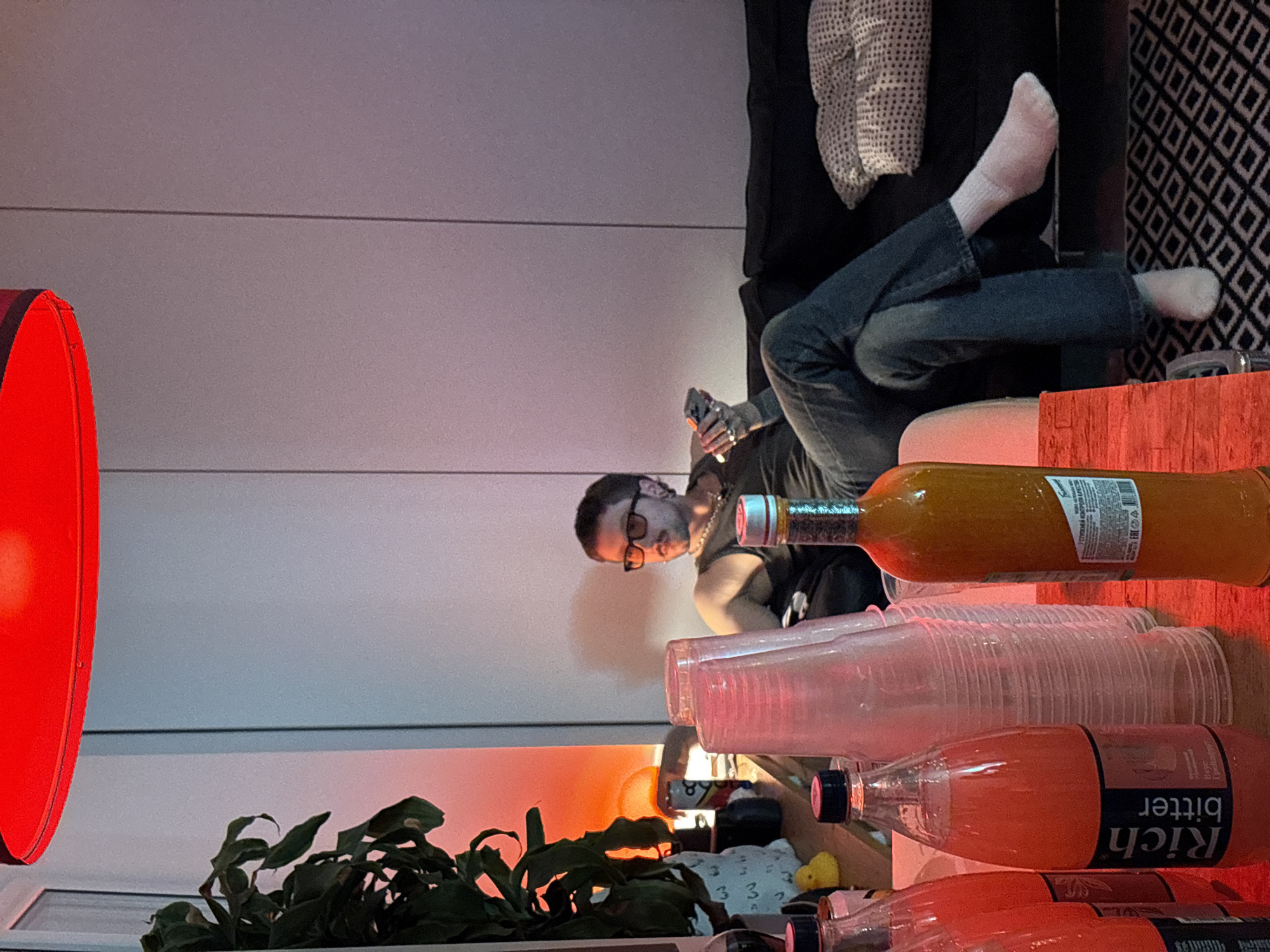 The image shows a casually arranged scene, likely indoors. A man wearing glasses and a t-shirt is visible, looking at or operating a mobile device. He is seated on a sofa with his legs extended, and the sofa is positioned against a wall with a textured pattern. In the foreground, there's a stack of clear plastic cups, several bottles of what appear to be beverages, and a potted plant with green leaves. A large red lamp is positioned on the left side of the frame, casting a strong red light. The image appears to be taken with a slight angle, and there are elements that may suggest a party or gathering setting. The location in the city of Moscow, Russia, is not directly indicated in the image, and requires additional information or context.