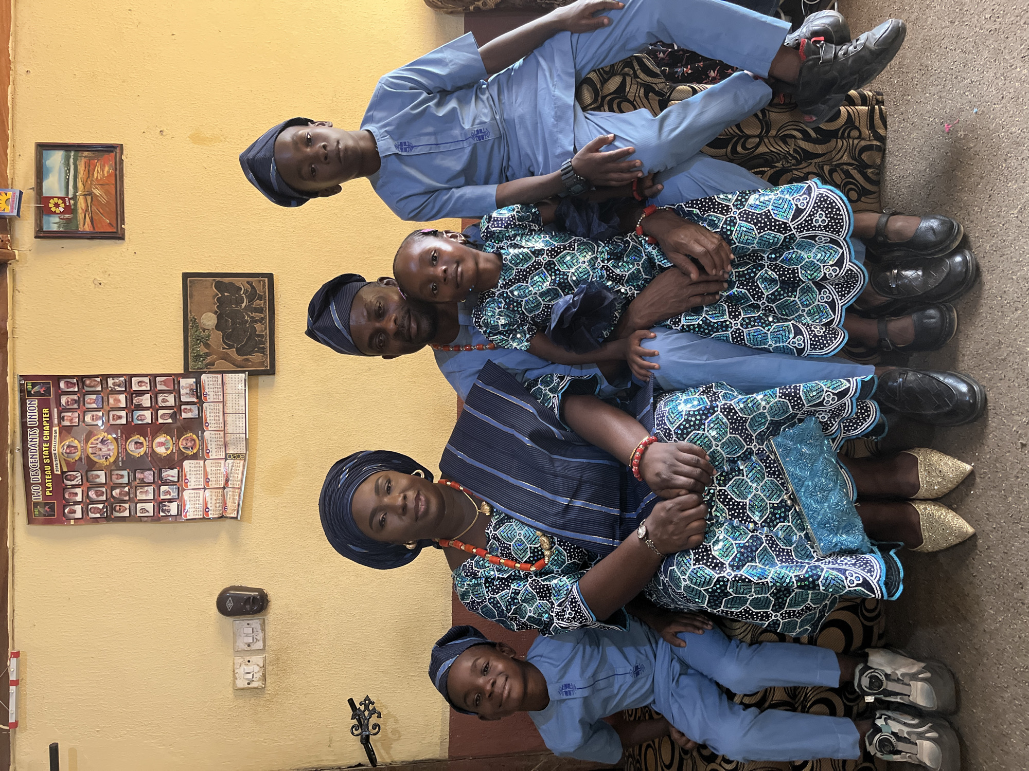 The image shows a family portrait taken in a room with yellow walls. A woman in an ornate blue and green dress and headwrap sits centrally, adorned with a red beaded necklace and matching bracelets. To her right sits a girl in a matching dress and a man in a blue outfit and hat. Two boys wearing blue outfits are positioned above and below. The room features framed artwork, light switches, and a calendar on the wall. The location is Rafin Bauna, Nigeria.