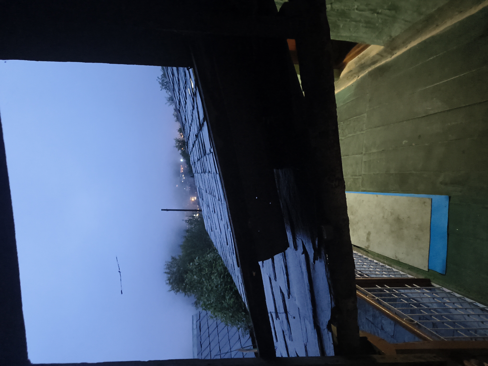 The image depicts a view from a covered porch or balcony overlooking a misty, twilight landscape with slate roofs. In the foreground, a dark wooden beam runs horizontally, obscuring the view below. Beyond the beam, slate-tiled roofs are visible, some appearing wet. Trees and buildings with glowing lights are discernible through the fog in the middle ground, suggesting a village or town. The sky is a pale, hazy blue, with a faint, dark linear object, possibly a distant bird or debris, visible in the upper left quadrant. The area below the porch appears to be a walkway or deck with a green surface and two mats, one blue and one beige. A metal railing runs along the left side of the walkway. The overall atmosphere is dim and slightly melancholic, indicative of early morning or late evening in a humid or misty environment. No people are visible in the image, and no specific location within the city of Haini, India, can be identified.
