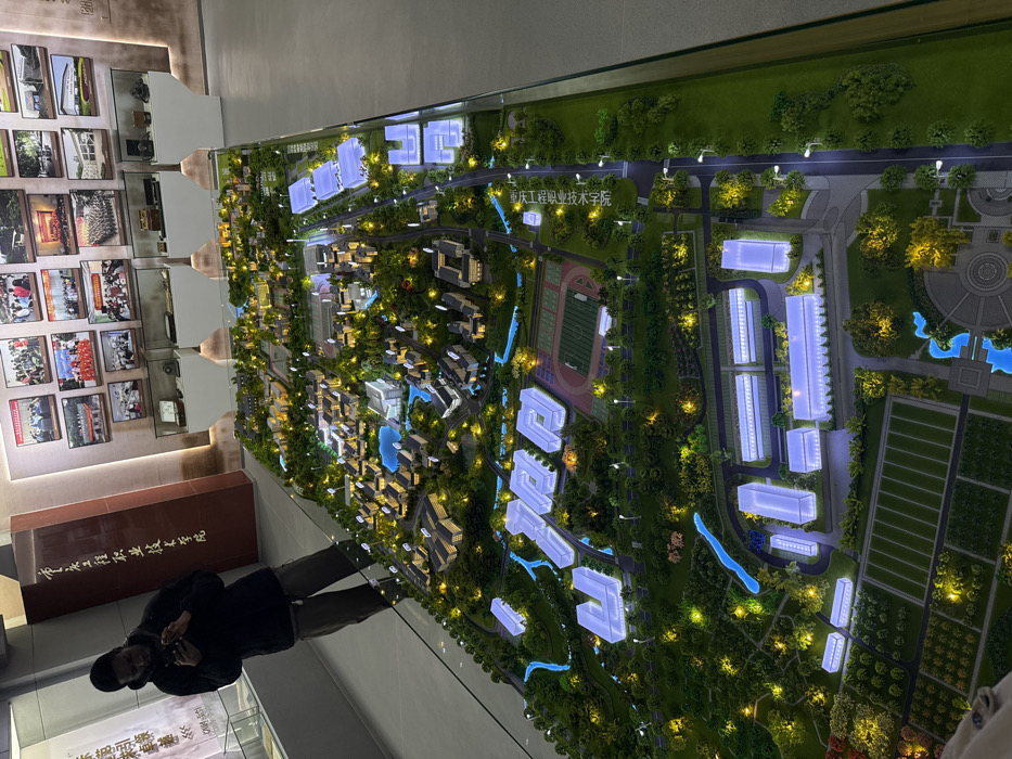 An indoor exhibition space, identified as Chongqing Jiangjin Vocational and Technical College in Jiangjin, China, by Chinese characters on a brown pillar, is depicted. The central subject is a large, illuminated architectural model of a college campus, displayed horizontally on a glass-topped surface. The model features numerous miniature buildings, some internally lit with white and blue light, surrounded by green landscaping with miniature trees, functional miniature streetlights, pathways, and blue-illuminated water features. An athletic track and field are visible within the model. A male individual, wearing a dark jacket and dark pants, stands to the left of the model, observing a smartphone. The background wall is adorned with multiple framed photographs of groups and campus scenes, positioned above several glass display cases with indiscernible contents. The floor is a light, reflective material.