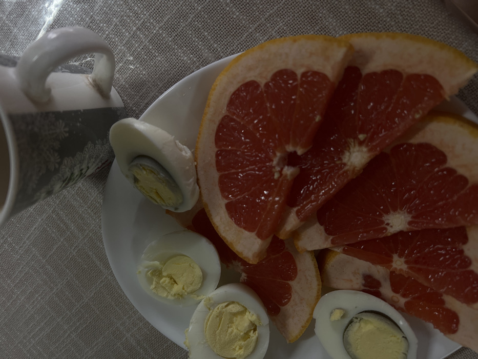 The image displays a close-up, overhead view of a meal placed on a light-colored, textured fabric surface. A white plate occupies the lower and central portions of the frame, holding several food items. On the plate are numerous crescent-shaped slices of red grapefruit, arranged somewhat overlapping. Alongside the grapefruit are four halves of hard-boiled eggs, each exposing a firm yellow yolk, with varying levels of irregularity in their cuts. A ceramic mug, partially visible in the upper right, features a dark background with light, possibly snowflake-like, patterns and contains a dark beverage. A bright highlight reflects off the mug's handle and extends onto the fabric surface, indicating a light source from the upper right. The environment details for the city of Leninakan, Russia, are not discernable from the visual information provided in this image.