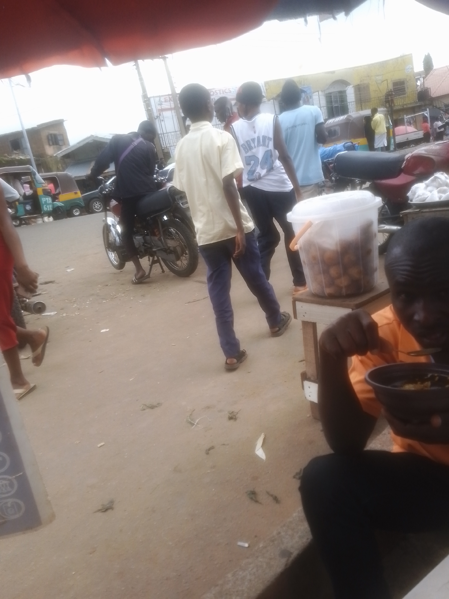 The image depicts a busy outdoor scene in Gwong, Nigeria. In the foreground, a person in an orange shirt is eating from a dark bowl with a spoon. To their right, a large white plastic container filled with food items, likely snacks or fritters, sits on a wooden table. Behind them, several people are visible. One person in a yellow shirt walks away from the camera, while another in a white basketball jersey with "BRYANT 24" on it and a person in a light blue t-shirt are walking in the same direction. A person is seated on a motorcycle, wearing a dark jacket with a purple strap. In the background, buildings, including a yellow structure, are visible, suggesting a marketplace or a street with shops. The presence of a tricycle, a common mode of transport in Nigeria, further reinforces the setting. The lighting suggests it is daytime, possibly overcast given the bright, diffused light. The overall impression is one of everyday activity in a Nigerian town.