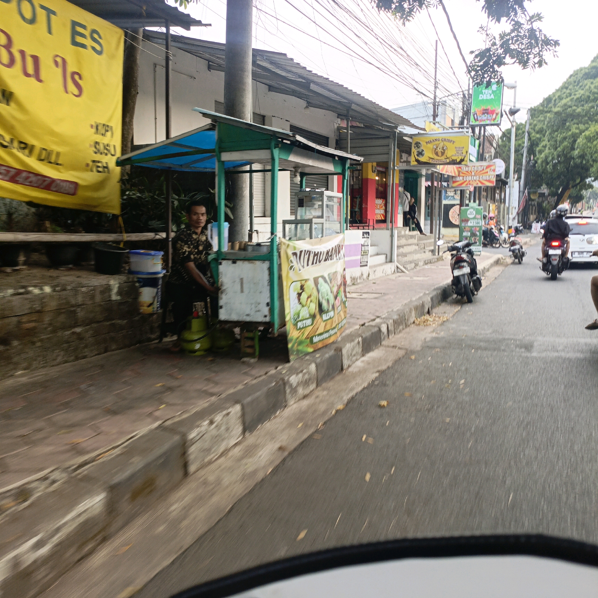 The image depicts a street scene in Salatiga, Indonesia, likely near a roadside food stall. A man in a dark patterned shirt and pants is seated behind a small food cart with a blue and green awning. Two green gas cylinders are positioned next to him. A yellow banner with Indonesian text, including "OT ES Bu Is" and "SARI DLL", is visible to the left of the food cart. Further down the street, other businesses with signage can be seen, including one with a yellow sign advertising "PISANG CRISPY". A motorcycle is parked in front of the food cart, and several other motorcycles and a car are present on the street. The road is asphalt, with a concrete curb and sidewalk. Trees are visible in the background, and the sky is overcast.
