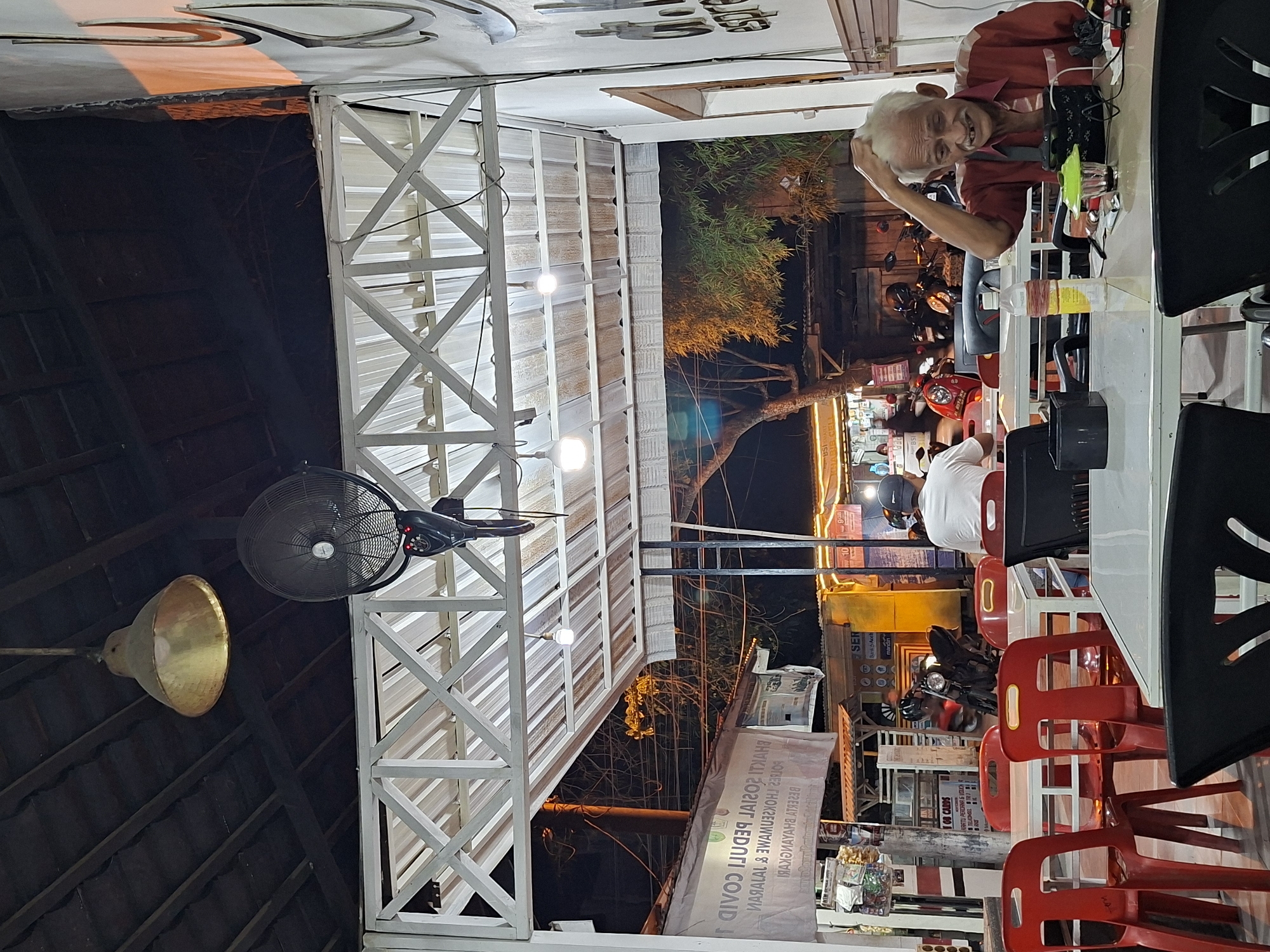 The image depicts a nighttime scene in what appears to be an outdoor dining area, potentially in Lhokseumawe, Indonesia. An elderly man with white hair is seated at a white table, looking at the camera and smiling. The seating consists of black and red plastic chairs. A fan is fixed to a white, cross-patterned support structure above the dining area. Various motorbikes are parked nearby, and a building with illuminated signage is visible in the background. The lighting is a mix of artificial light from lamps and signage, as well as natural ambient light. A sign with Indonesian text related to Covid is partially visible.