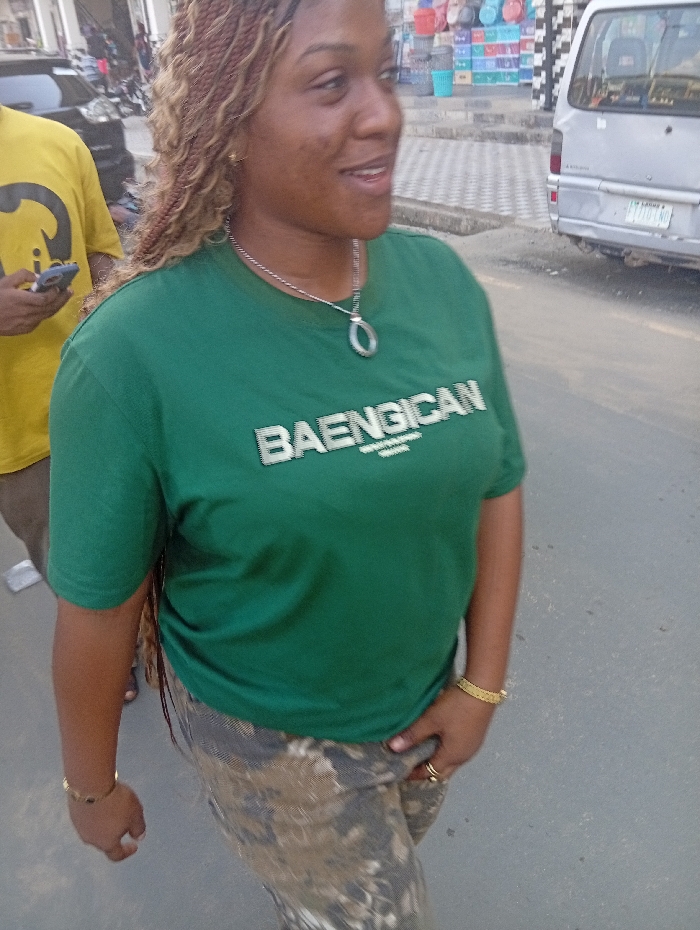 A woman with light brown braided hair wears a green "BAENGICAN" t-shirt, patterned pants, and gold jewelry. She has a silver necklace with a circular pendant. A man in a yellow shirt partially obstructs the left of the frame, holding a phone. A silver van is visible on the right, parked on a paved road. In the background are storefronts with assorted items on display.