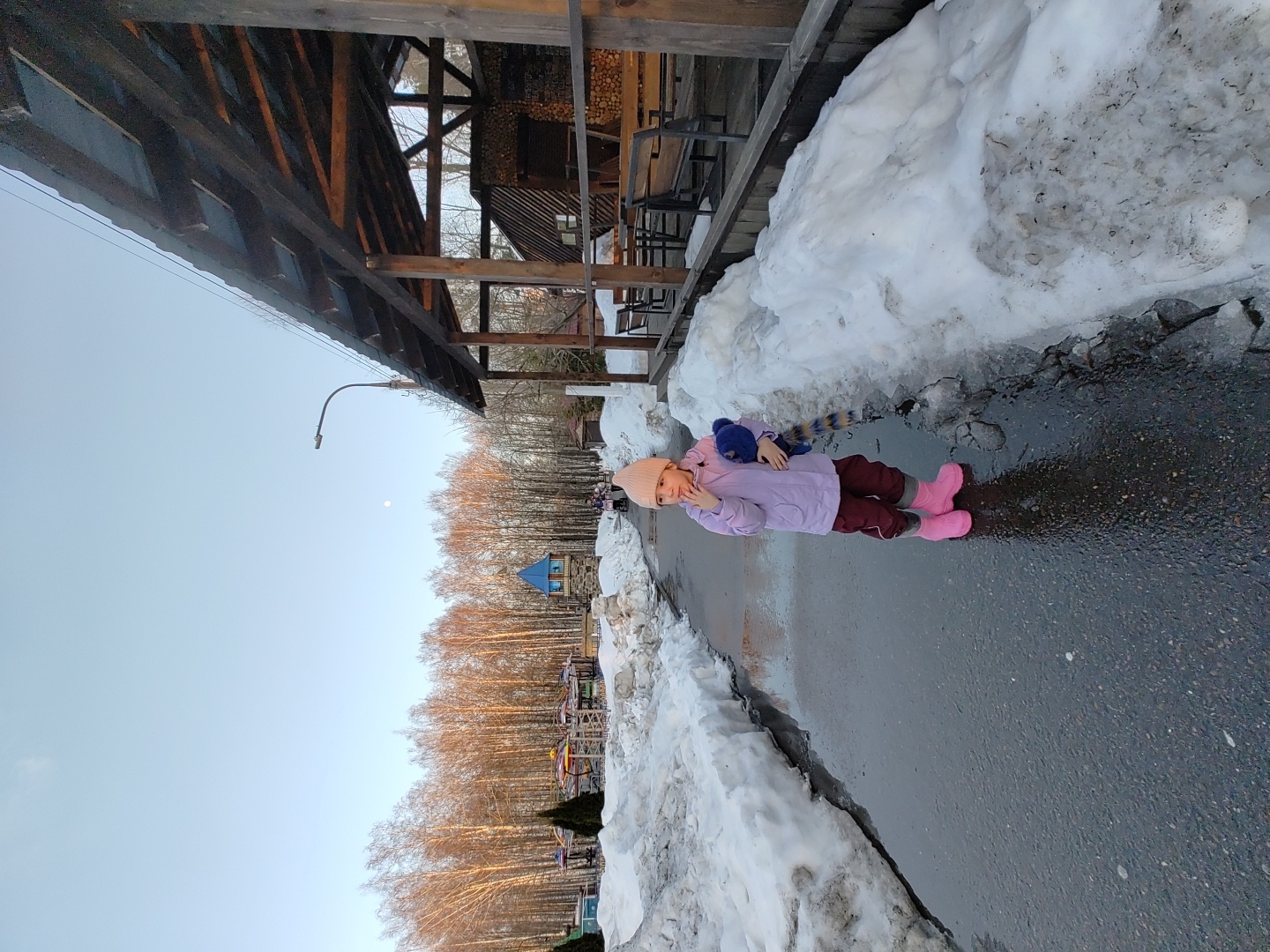 A young child wearing a pink hat, light purple jacket, maroon pants, and pink boots is standing on a wet asphalt path. She is holding a stuffed animal with a striped tail. To the left, there is a large pile of snow. In the background, there are bare trees and a playground with a blue roof. In the foreground, there is a wooden structure with seating. The location is Chaikovsky, Russia.