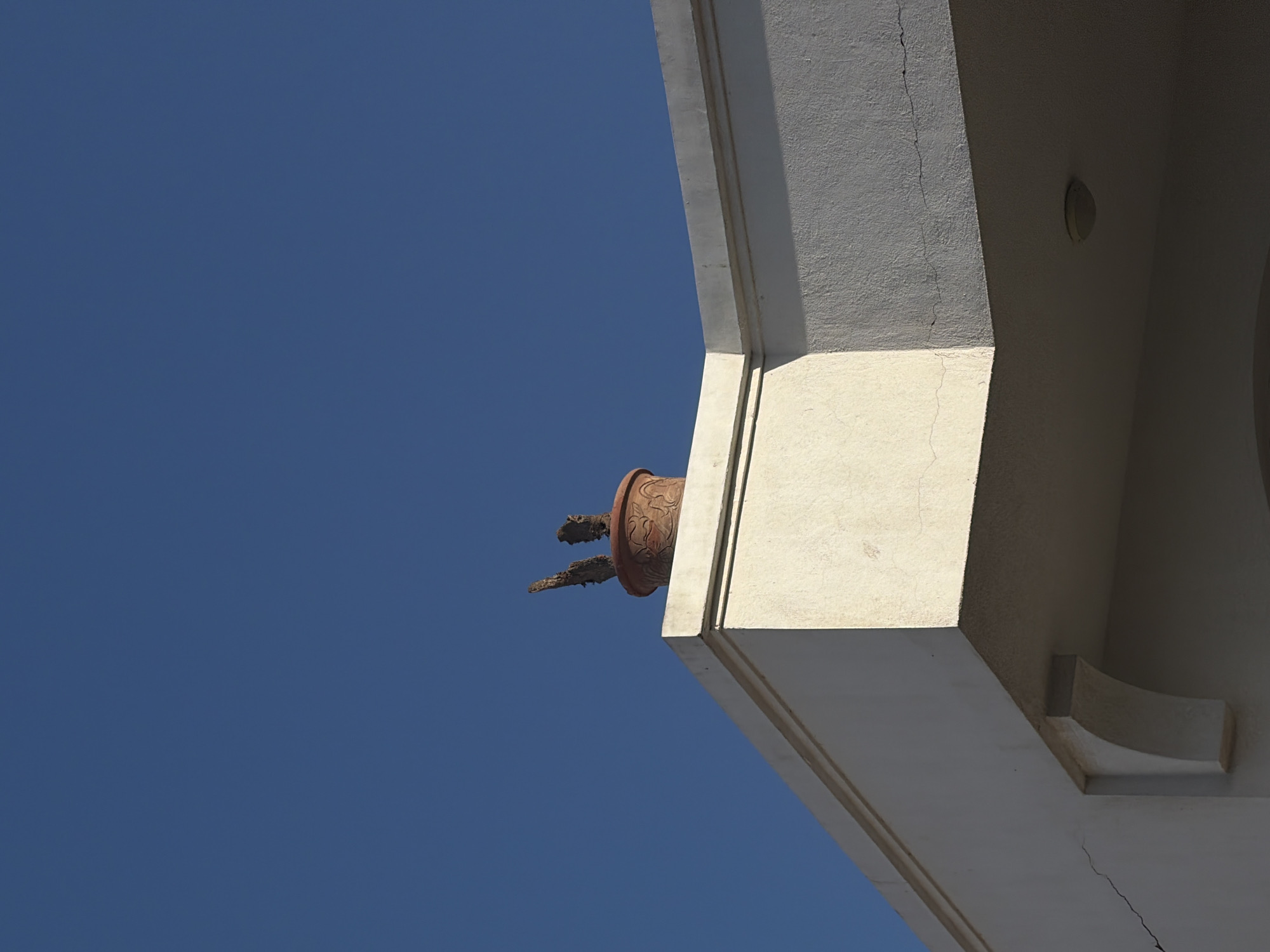 The image depicts a terracotta pot with two dried branches inside, placed on the edge of a building's roof against a clear blue sky. The building appears to be a white, stucco structure with architectural detailing, including an overhang and a decorative bracket. The pot has an intricate, embossed pattern around its rim. The scene is well-lit by sunlight, casting distinct shadows. The overall impression is one of stillness and a minimalist aesthetic, potentially located in a city like Sharm El Sheikh, Egypt, where such architectural styles and clear skies are common. There are no people or animals visible in the frame.