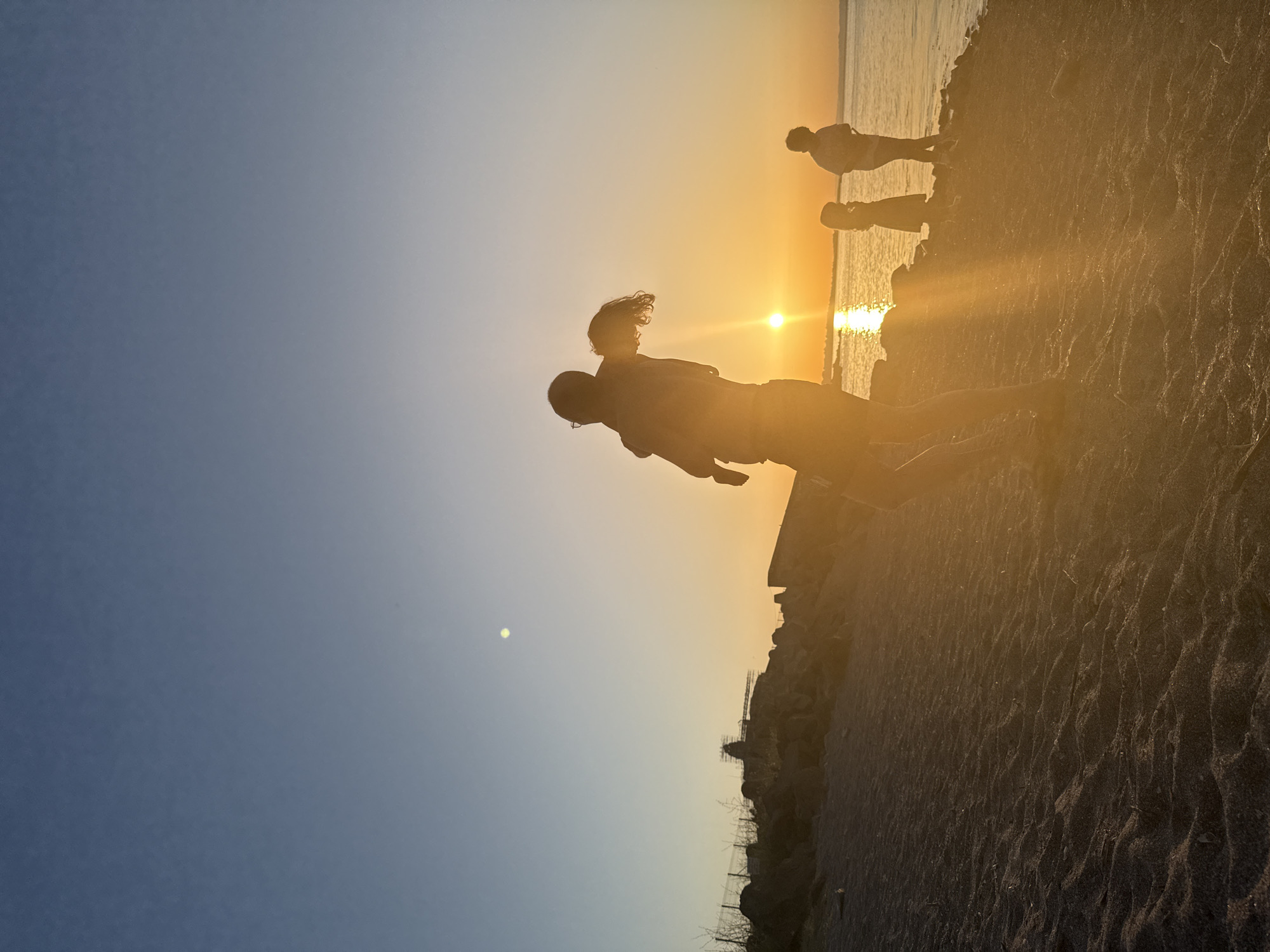A silhouetted family is enjoying the sunset at a beach in Sozopol, Bulgaria. The scene is illuminated by the warm glow of the setting sun, which is low on the horizon and casting long shadows. A man is holding a child on his shoulders, while two other children stand nearby. The water is calm, reflecting the golden hues of the sky. The beach is covered in sand, with some rocks and vegetation visible along the shoreline. The overall mood is peaceful and joyous, capturing a moment of family connection during a beautiful sunset.