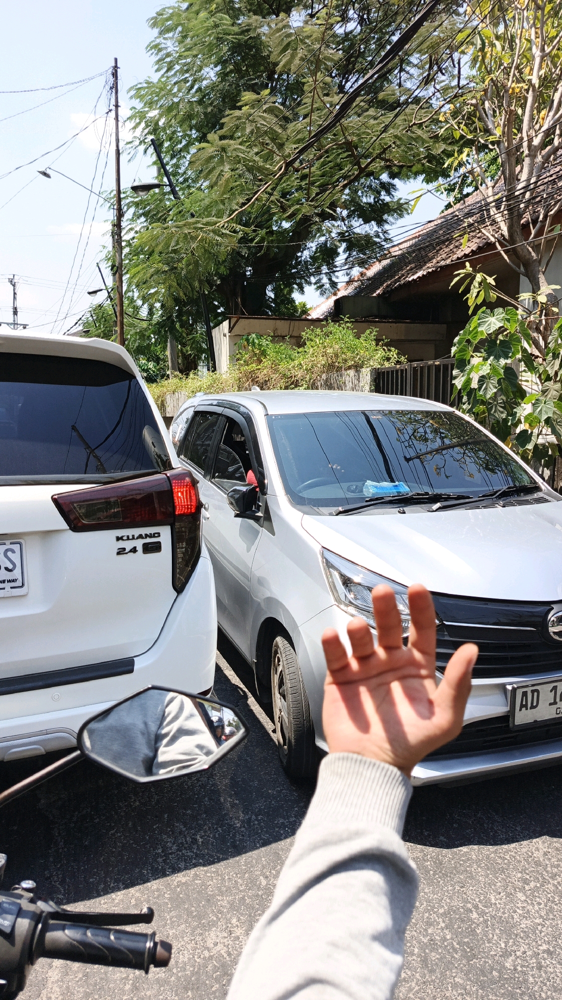 In Surakarta, Indonesia, two cars are parked closely on a street under a bright sky. A white car, identified by the "KIJANG 24" emblem, is on the left, and a silver car with license plate "AD 14" is on the right. The silver car's windshield displays a blue item. A person's hand and arm, clad in a gray sweater, extend into the foreground, obscuring a portion of the silver car's front. The view suggests the image is taken from a motorcycle, as indicated by the handlebars and a rearview mirror reflecting the person in the gray sweater and a glimpse of a motorcycle. Trees and power lines are visible in the background, alongside a residential building. The sunlight casts distinct shadows on the asphalt.
