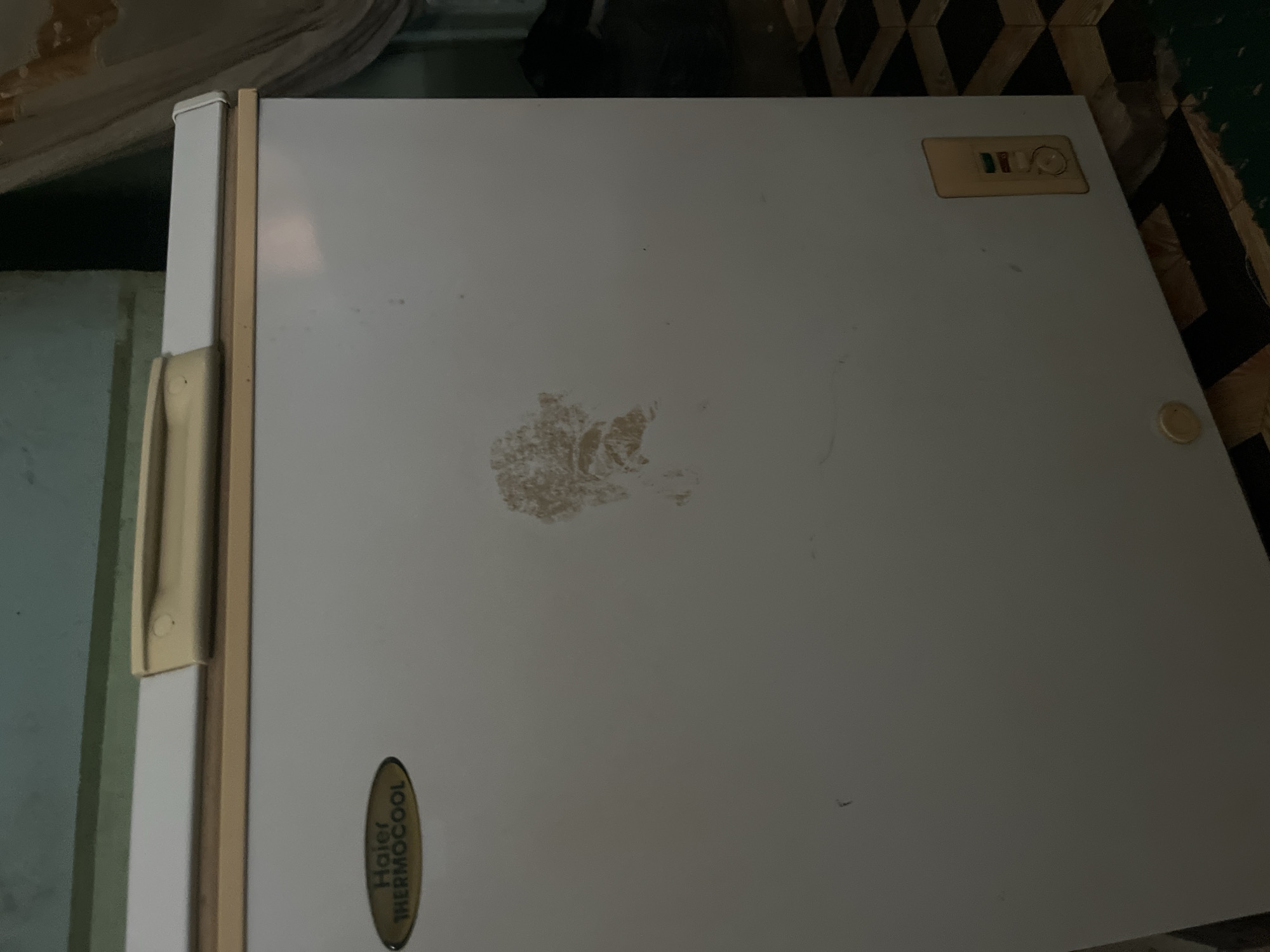 The image shows the exterior of a white refrigerator with a gold handle on the left side. On the top right of the refrigerator door, there is a gold-colored badge with the text "BITLO" and some colorful indicators, possibly representing temperature settings. Towards the bottom left, there is an oval-shaped badge with the text "Haier THERMOCOOL" in green and black lettering. There is a light brown stain or mark in the center of the refrigerator door. The refrigerator is set against a backdrop that includes a patterned rug or flooring and a wall. The lighting in the image is dim, suggesting it was taken indoors in low light conditions. The overall impression is of an older appliance in a home environment. The location is Jauro Yinu, Nigeria.