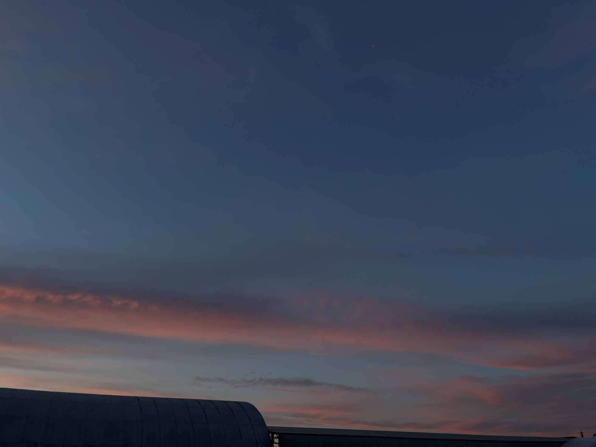 The image depicts a twilight sky over what appear to be industrial or agricultural structures in Novopokrovskaya, Russia. The sky transitions from a deep indigo-blue in the upper frame to lighter blue-gray hues, with distinct horizontal bands of stratiform clouds illuminated in soft pink and orange, consistent with a sunset. In the lower portion of the frame, two dark building silhouettes are visible. On the left, a large, curved, corrugated metal structure, possibly a quonset hut or hangar, occupies the lower-left. To its right, a lower, flatter roofline of another building extends along the horizon. Several small, faint, reddish-pink dots are present in the upper and mid-sky, consistent with lens flares or sensor artifacts. No individuals or discernible activities are present.