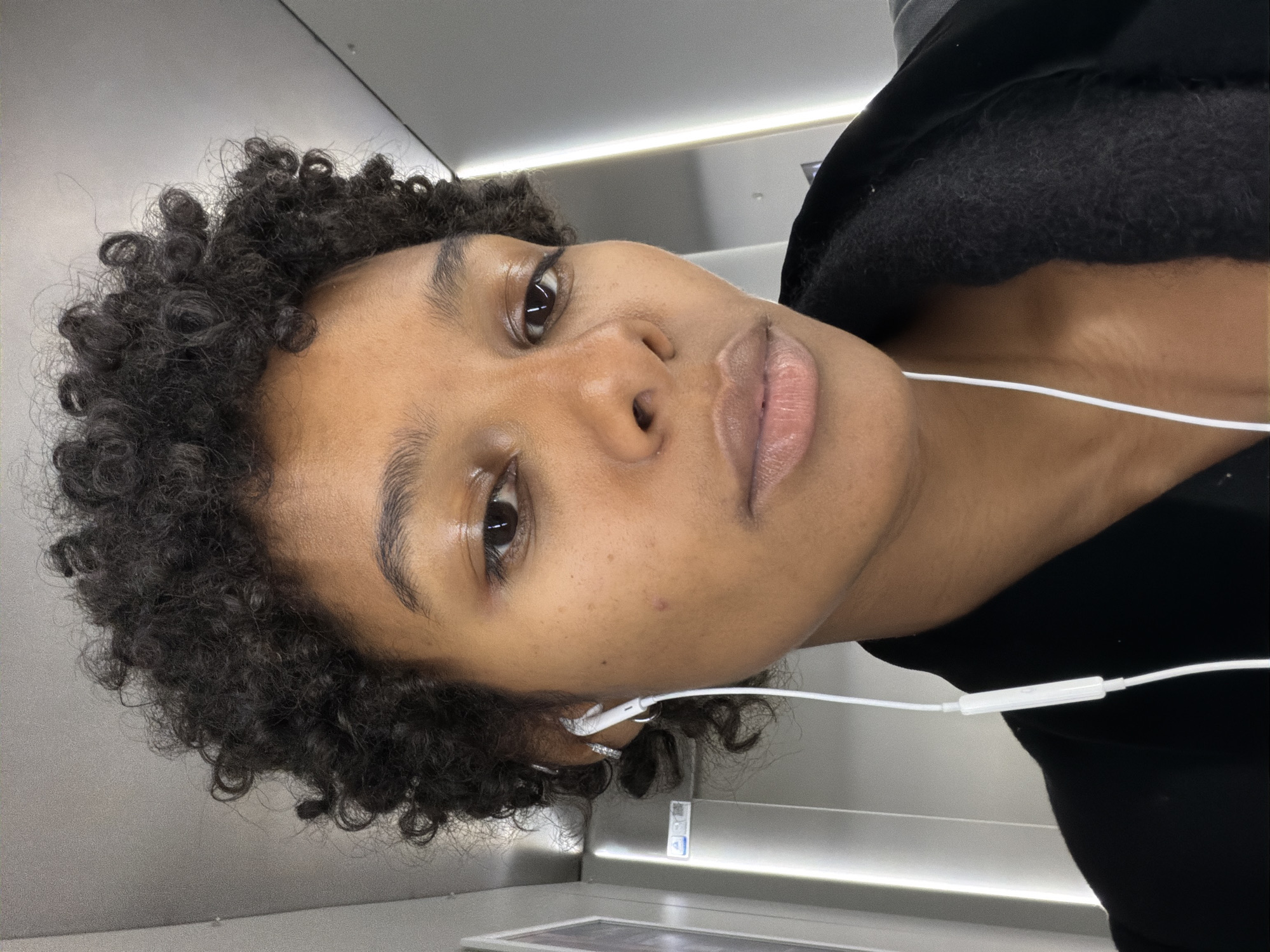 A close-up, head-and-shoulders shot shows a young Black woman with short, curly dark hair, wearing white earbuds and a black jacket. She is looking directly at the camera with a neutral expression. The background appears to be an indoor metallic hallway, possibly a train or subway car, with bright vertical lighting. There is no indication of the city of Bremen, Germany in the image.