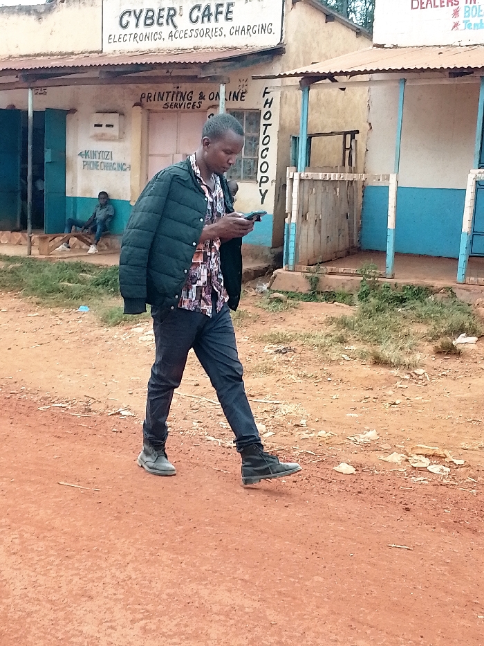 The image shows a man walking down a dirt road in Kivaywa, Kenya, located at latitude 0.562 and longitude 34.805. The man is wearing a dark green jacket, a multi-colored shirt, dark blue pants, and black shoes. He is looking at a mobile phone in his right hand. The road is red, and there are buildings on either side. One building has a sign that reads "Cyber Cafe, Electronics, Accessories, Charging." Another building has a sign that reads "Printing & Online Services." There is also a sign that reads "Photocopy." The man is walking towards the right side of the image. There is a small amount of vegetation on the sides of the road. There is a man sitting on a bench behind the person in the foreground. The man is wearing a dark blue jacket. The background is out of focus.