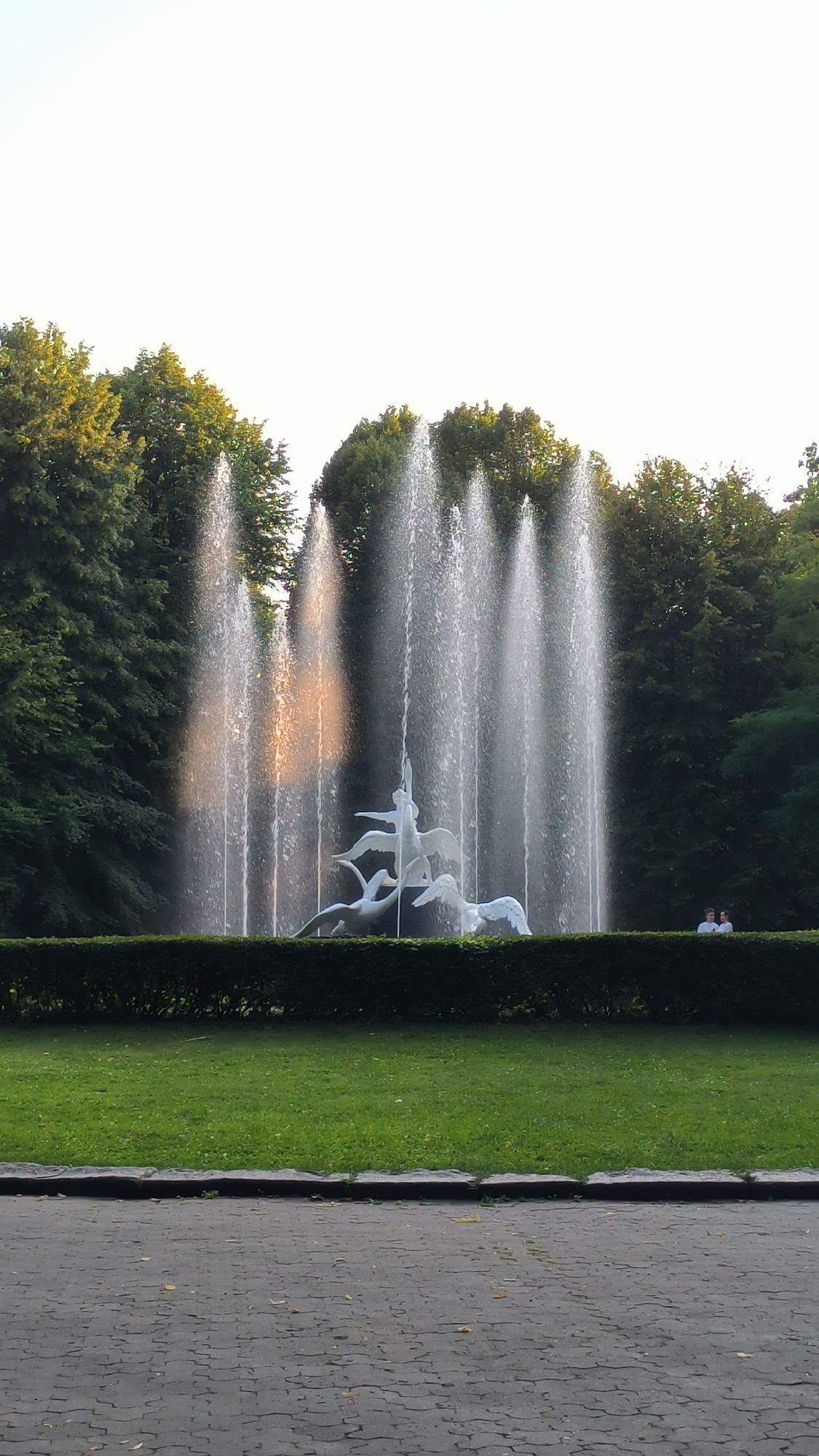 The image captures a picturesque fountain adorned with white sculptural figures of swans and humanoids, set against a backdrop of lush green trees. The fountain is actively spraying water, creating a dynamic visual effect. In the distance, two individuals are visible, observing the fountain. The foreground features a paved walkway, suggesting a public park or promenade setting. The overall scene conveys a serene and tranquil atmosphere. The fountain is located in Lviv, Ukraine.