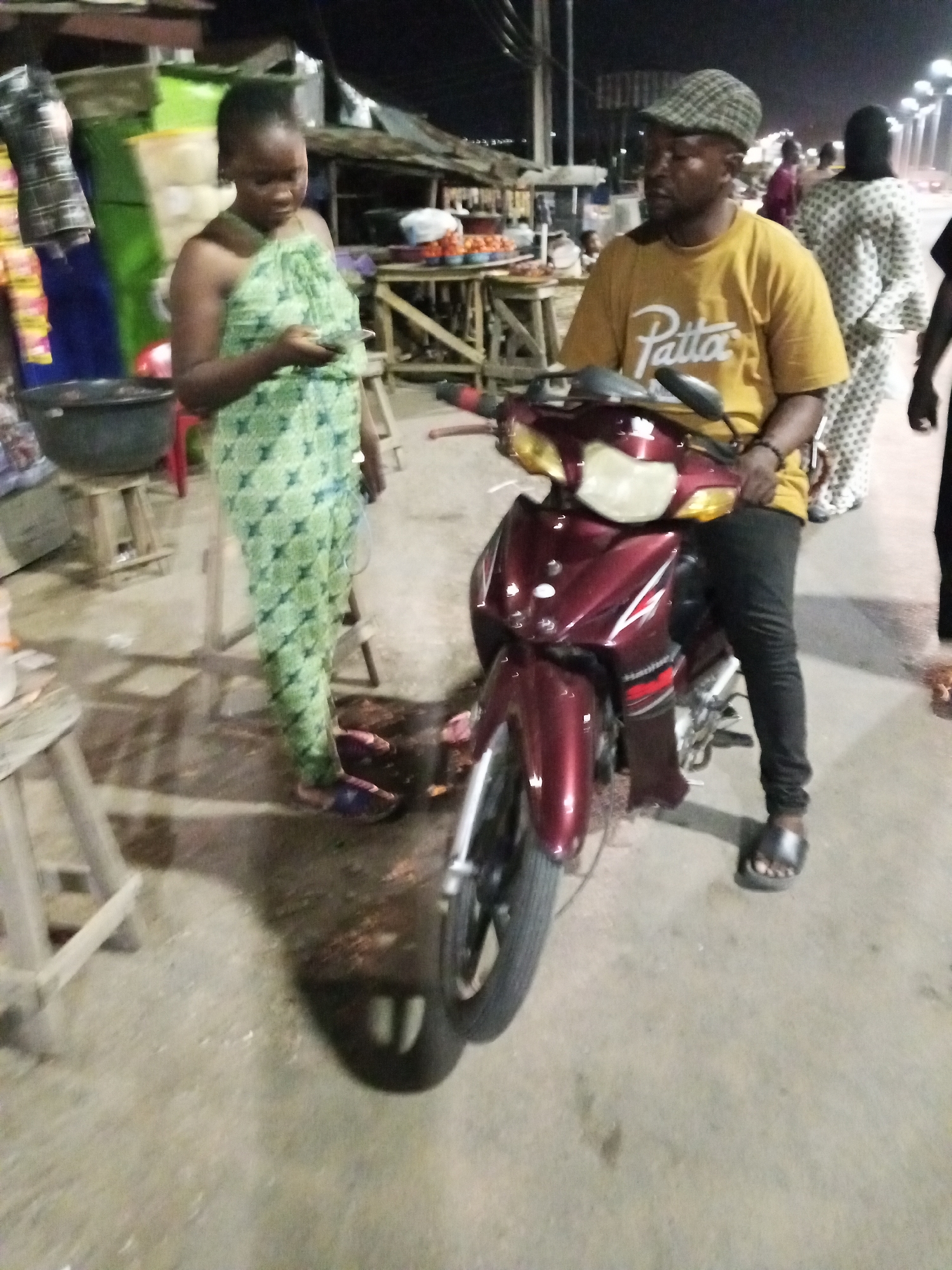 The image depicts a street scene in Adankolo, Nigeria, at night. A woman in a green patterned halter-style jumpsuit stands to the left, looking at a mobile phone. A man wearing a yellow "Patta" t-shirt and a patterned cap is sitting on a maroon motorcycle to the right, looking at the woman. The street is a gray concrete surface. Stalls with goods are in the background. Street lights are visible in the distance. Additional figures are walking along the street in the background.